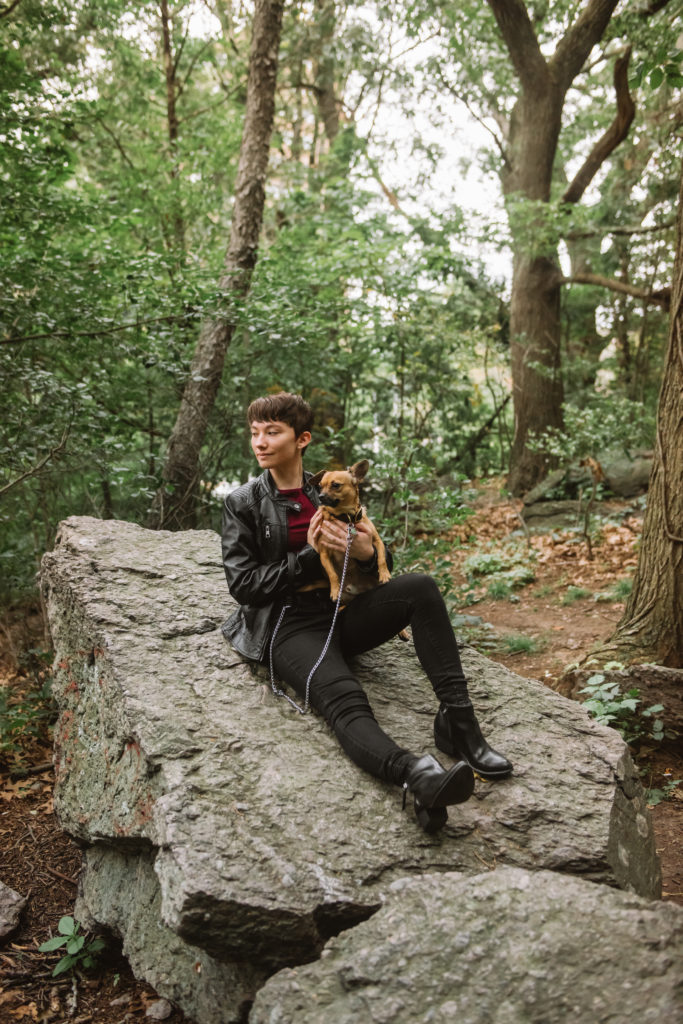 Danielle in a black leather jacket sitting on a large rock holding Milo. They are both looking off to their right.