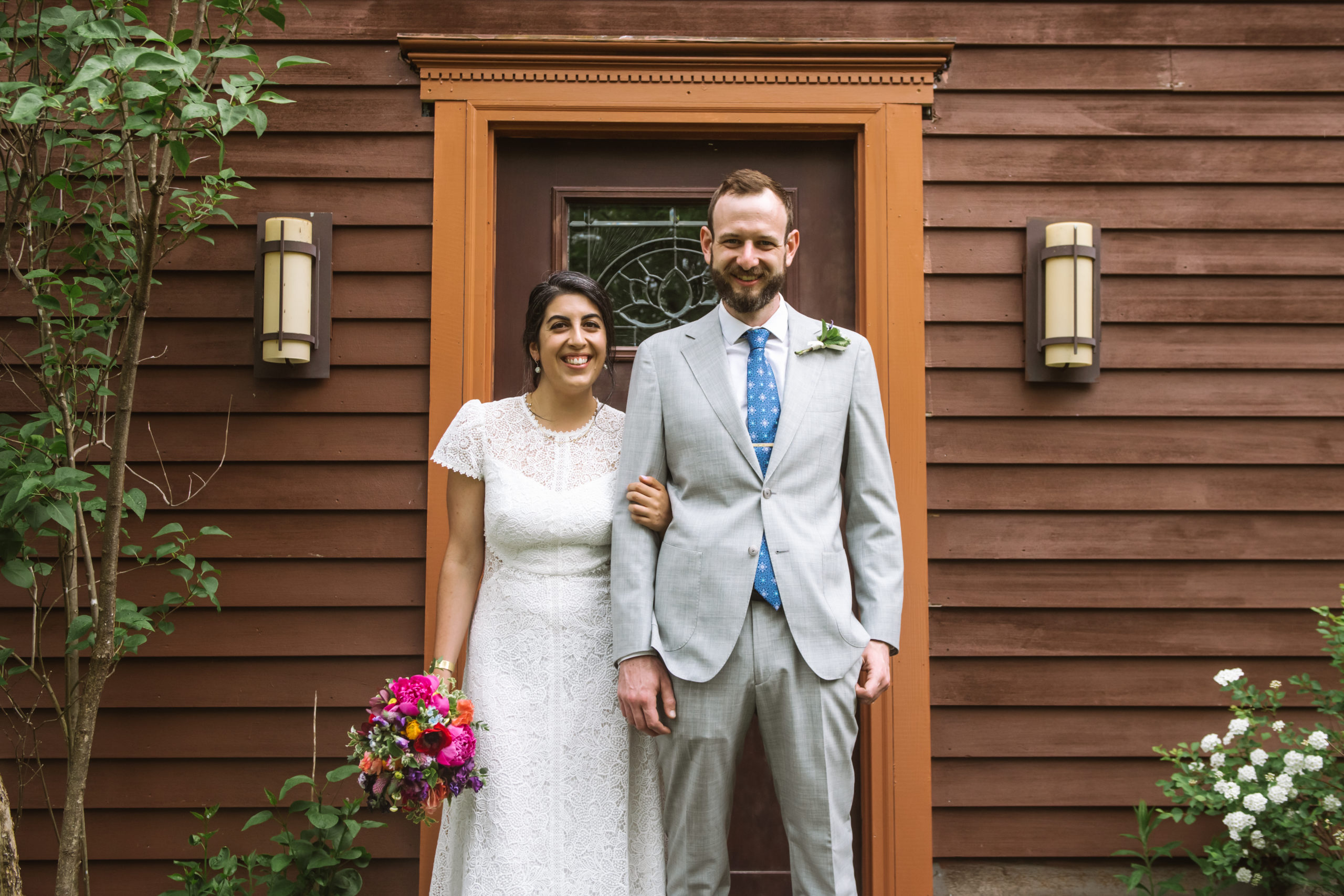 Bride and groom standing side by side, with the bride's left hand wrapped around the groom's right arm. They are both smiling, looking straight to the camera. The bride is holding her bouquet in her right hand and the groom has both hands relaxed at his sides. They are standing in front of a dark red wood house's front door with greenery and trees surrounding the facade.