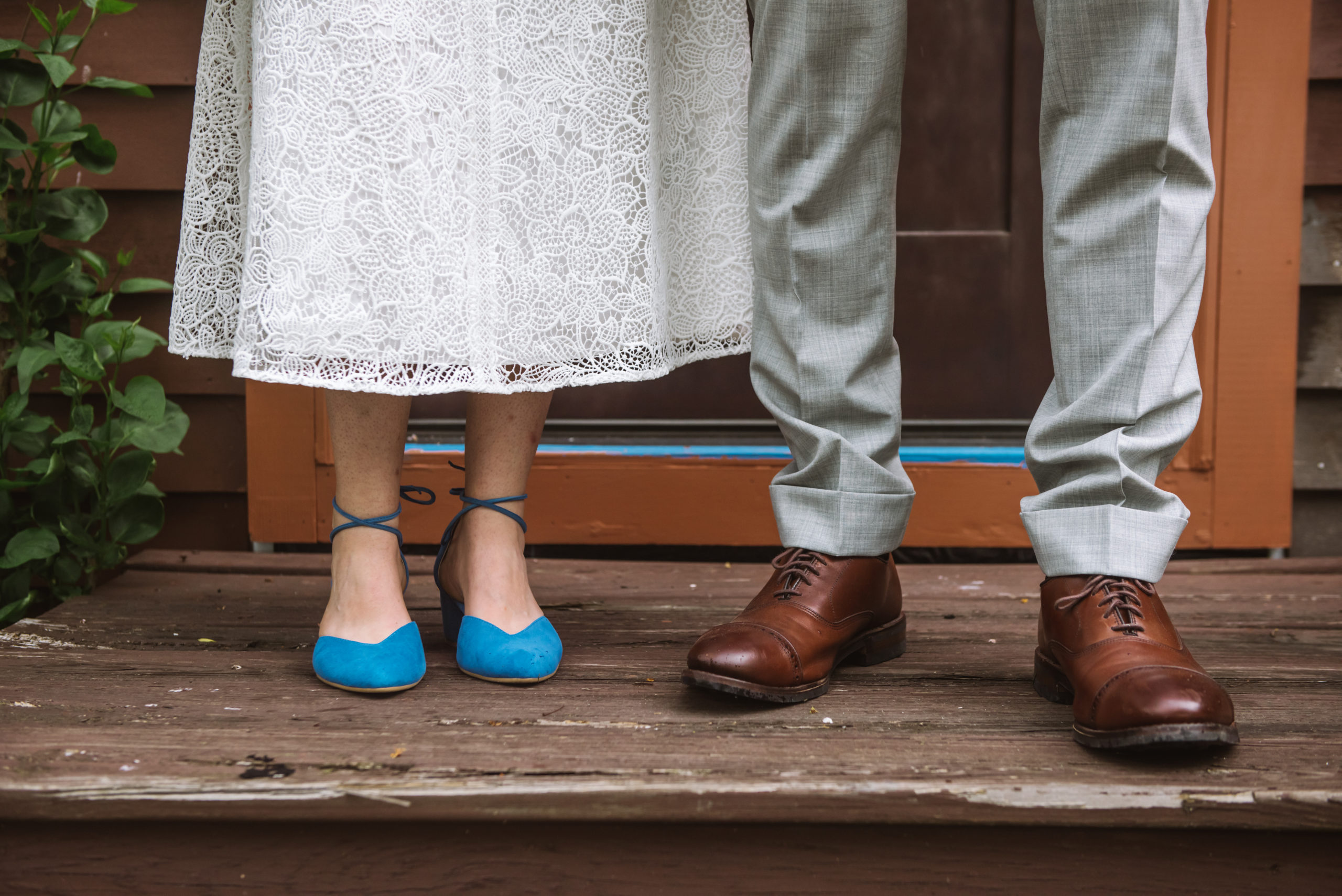 Focus on the bride and groom's feet and ankles. They are standing on a wooden stoop in front of the house's front door. The bride is wearing a white lace dress with blue strappy low heels. The groom is wearing brown dress shoes and light grey suit pants.