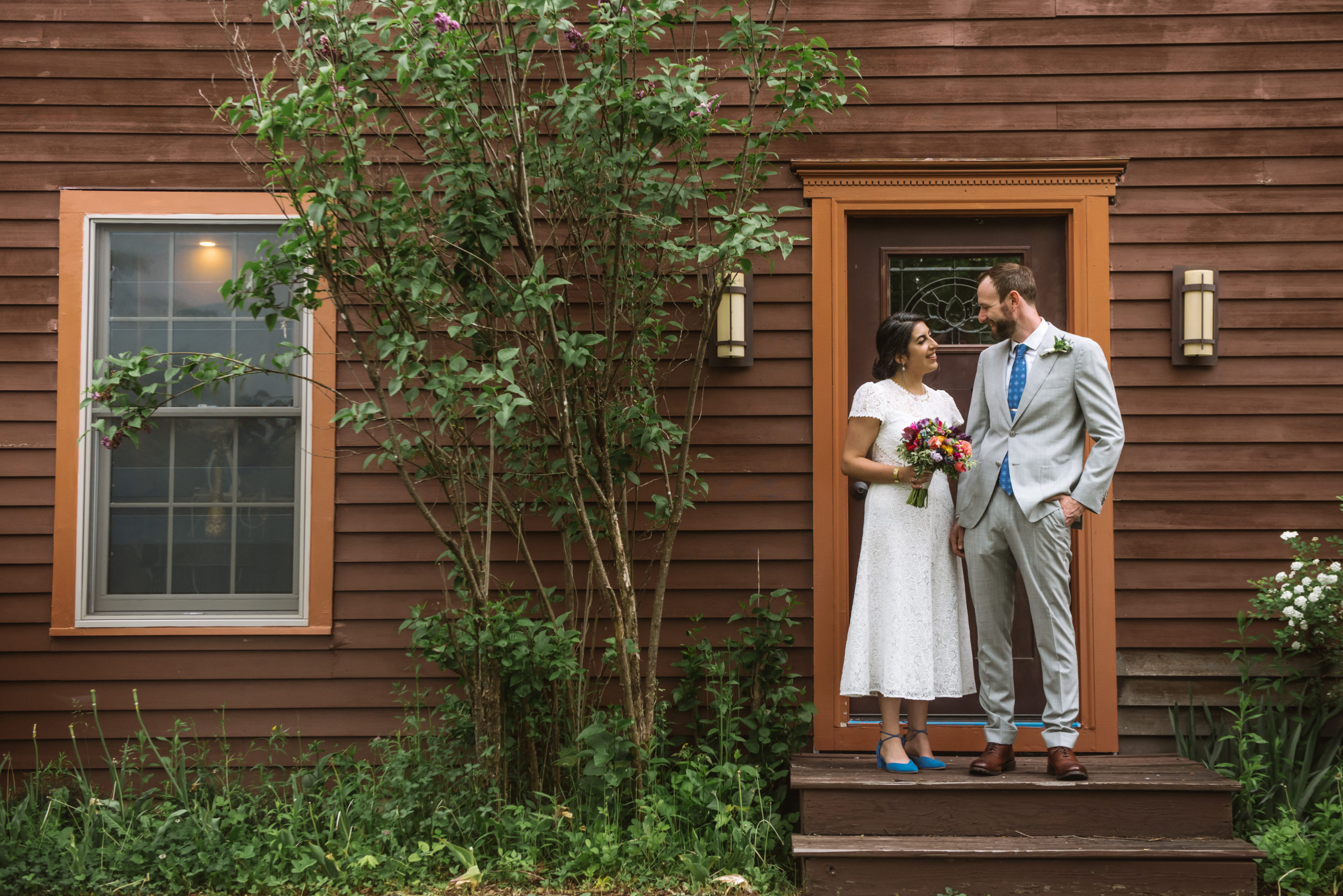 Bride and groom standing side by side, facing one another. They are both smiling. The bride is holding her bouquet in her right hand and the groom has his right hand relaxed at his side with his left hand in his pocket. They are standing in front of a dark red wood house's front door with greenery and trees surrounding the facade.