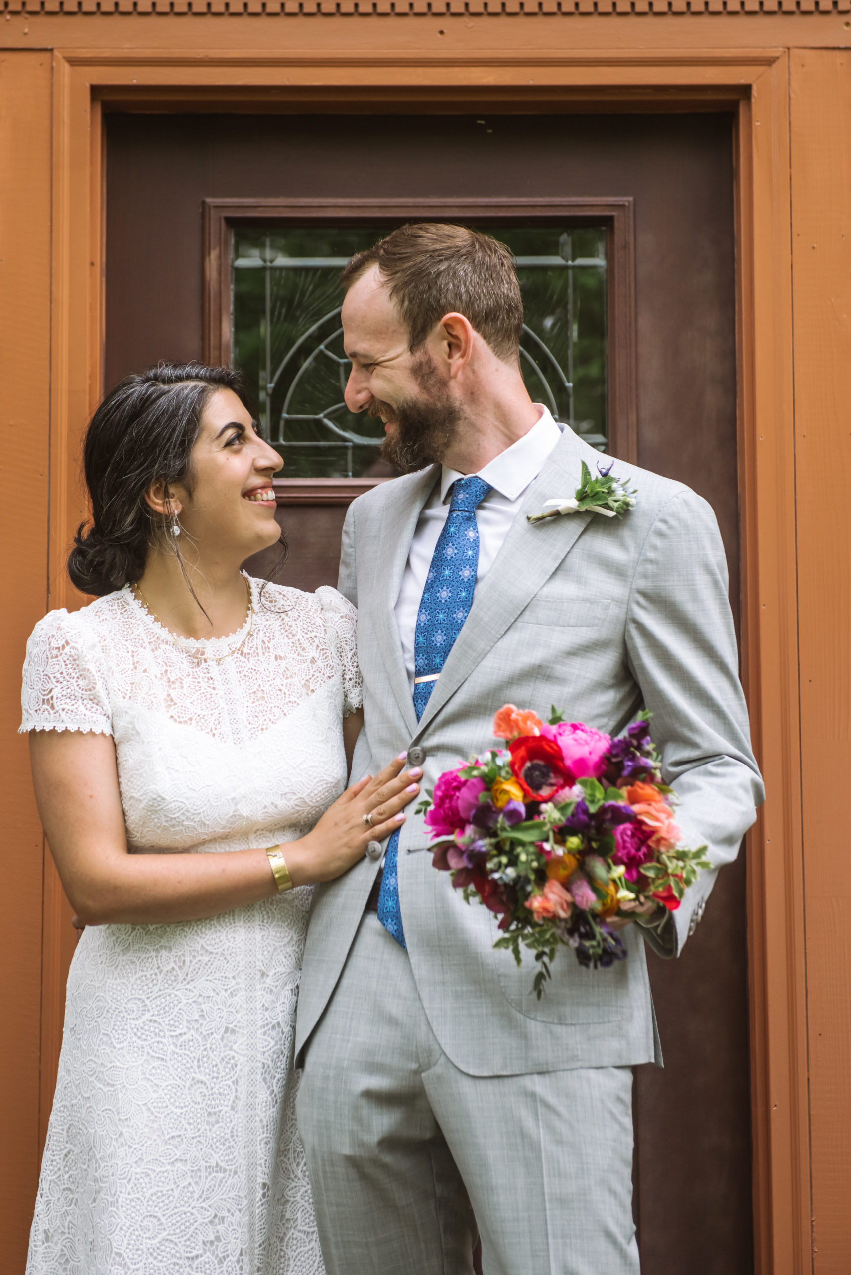 Bride and groom standing side by side, heads turned toward one another. They are both smiling. The bride is resting her right hand on her husband-to-be's waist. The groom is holding his wife-to-be's bouquet in his left hand. They are standing in front of a dark red wood house's front door with greenery and trees surrounding the facade.