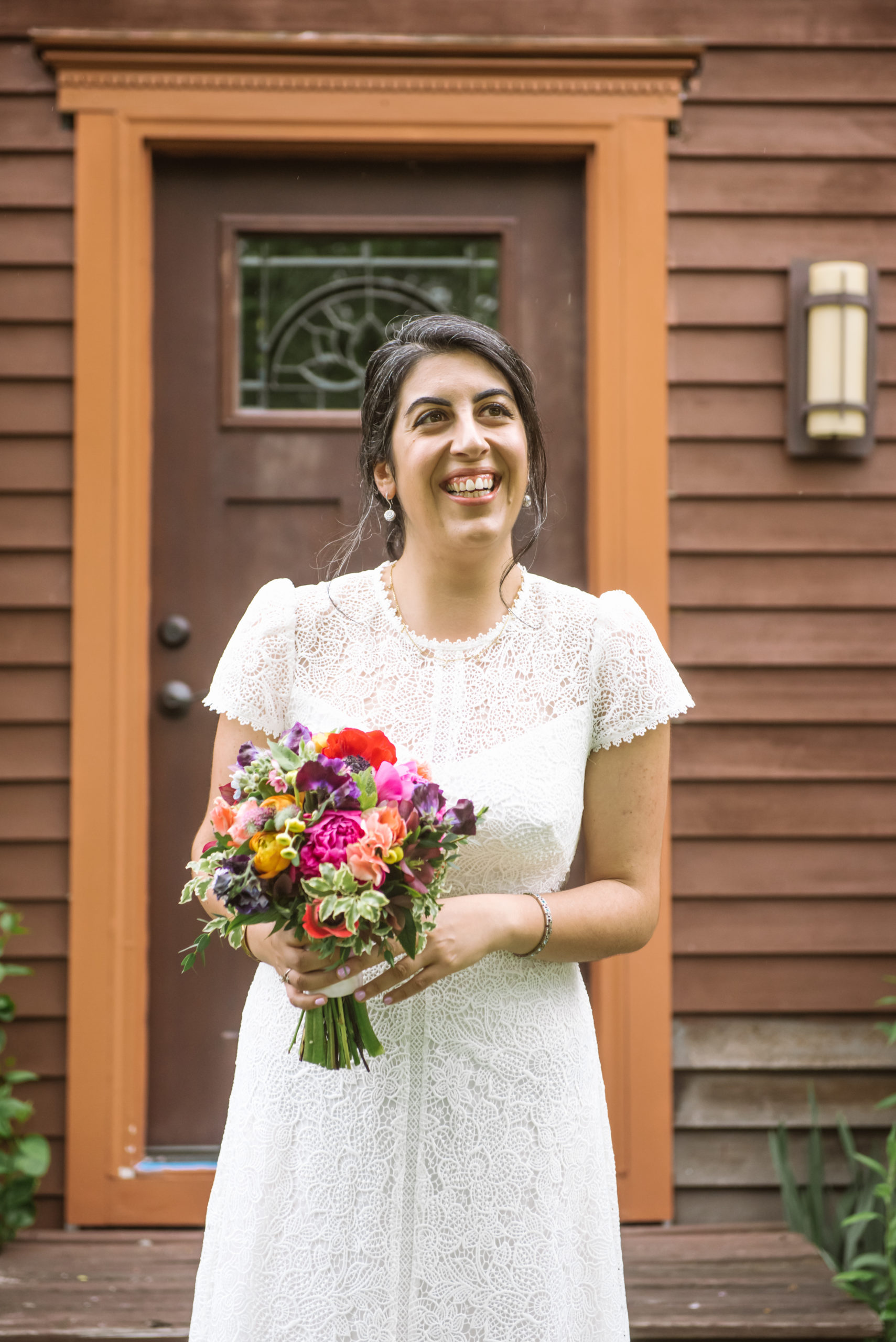 Bride holding her bouquet in her hands. She is wearing a white lace dress with short sleeves. She is standing in front of a dark red wood house's front door.