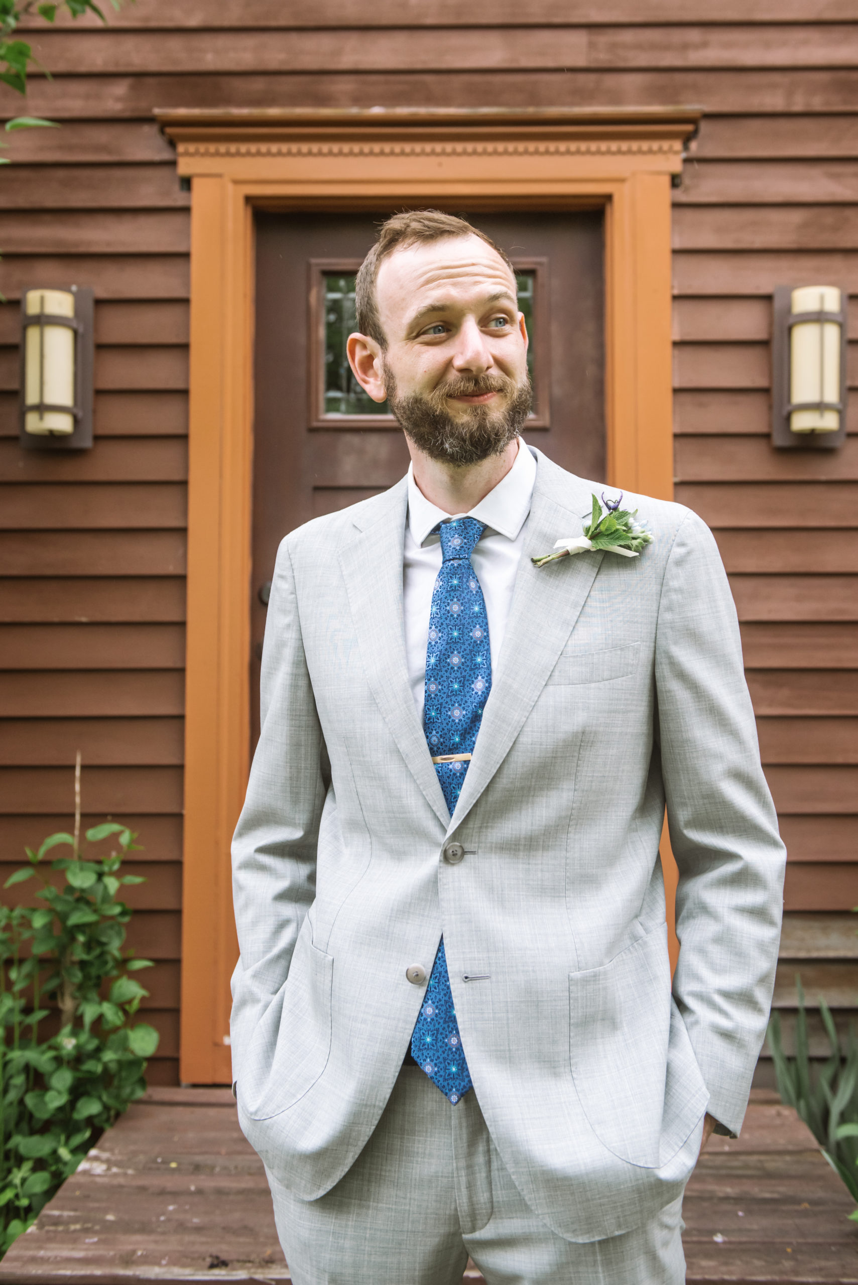 Groom wearing a light grey suit, blue patterned tie, gold tie clip, white collared shirt, and a boutonniere looking off to his left with a soft smile on his face. He is standing in front of a dark red wood house's front door with greenery and trees surrounding the facade.