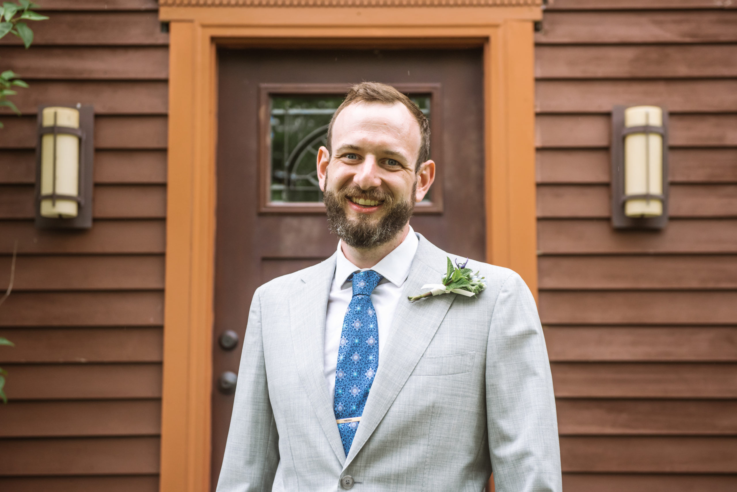 Groom wearing a light grey suit, blue patterned tie, gold tie clip, white collared shirt, and a boutonniere looking straight to camera and is smiling. He is standing in front of a dark red wood house's front door with greenery and trees surrounding the facade.
