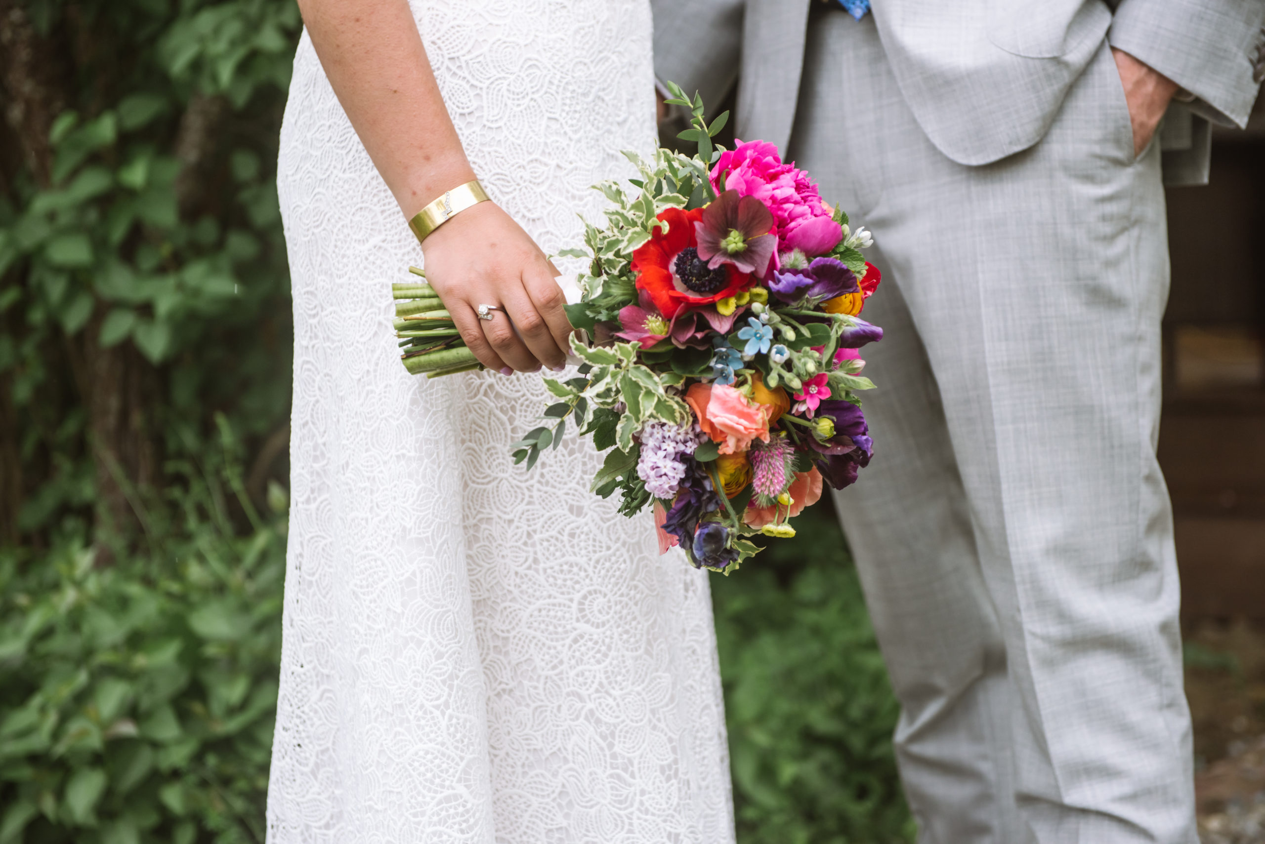 Close up photo of the bride's bouquet in her right hand. She is wearing her engagement ring (a family heirloom) on her ring finger and is wearing a bracelet. Bride and groom standing facing one another. The bride is holding her multi-colored bouquet in her right hand and the groom's left hand is in his pocket.