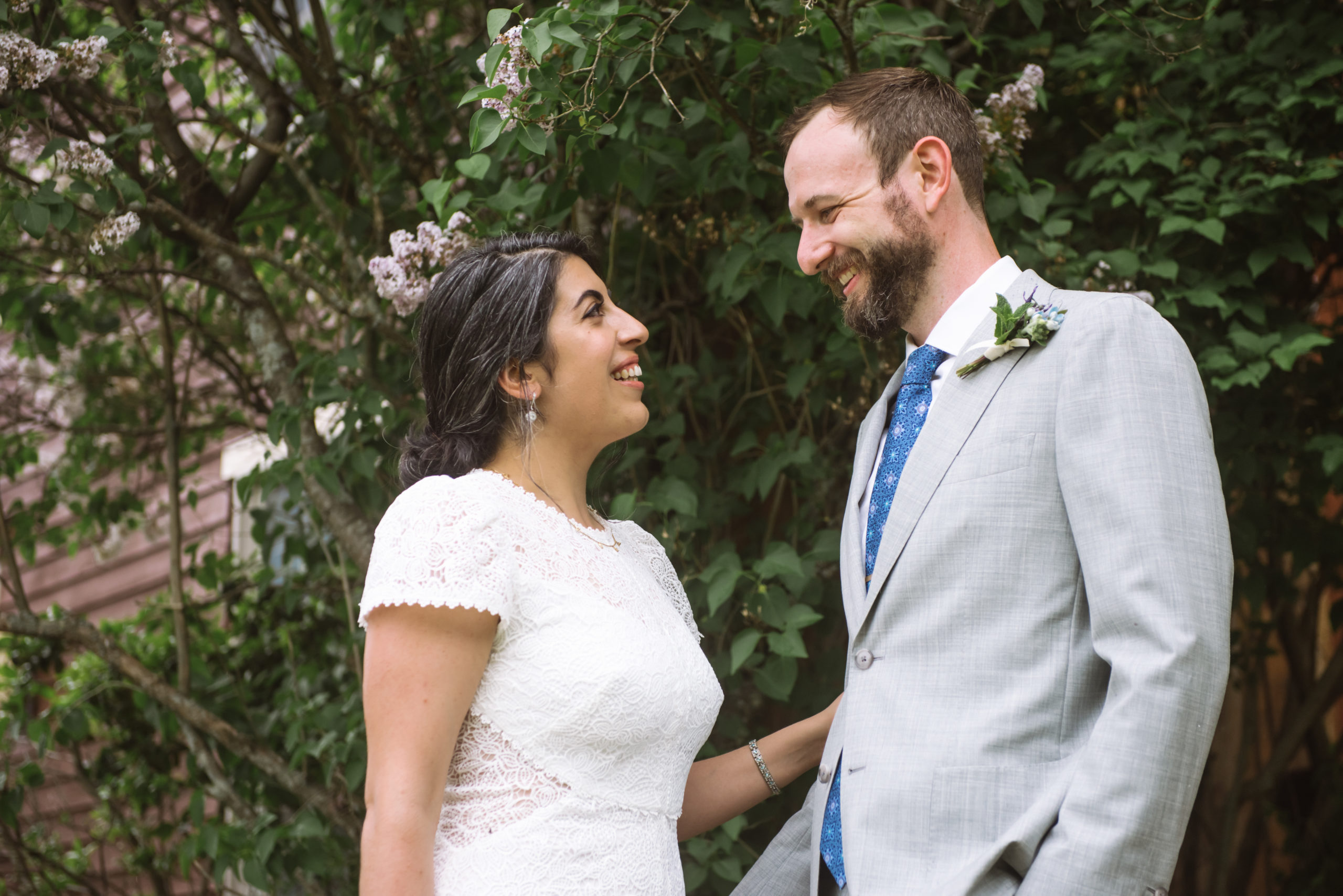 Bride and groom smiling at one another. One of each of their hands/arms is touching the other. They are standing in front of a tree with green leaves and white/purple flowers.