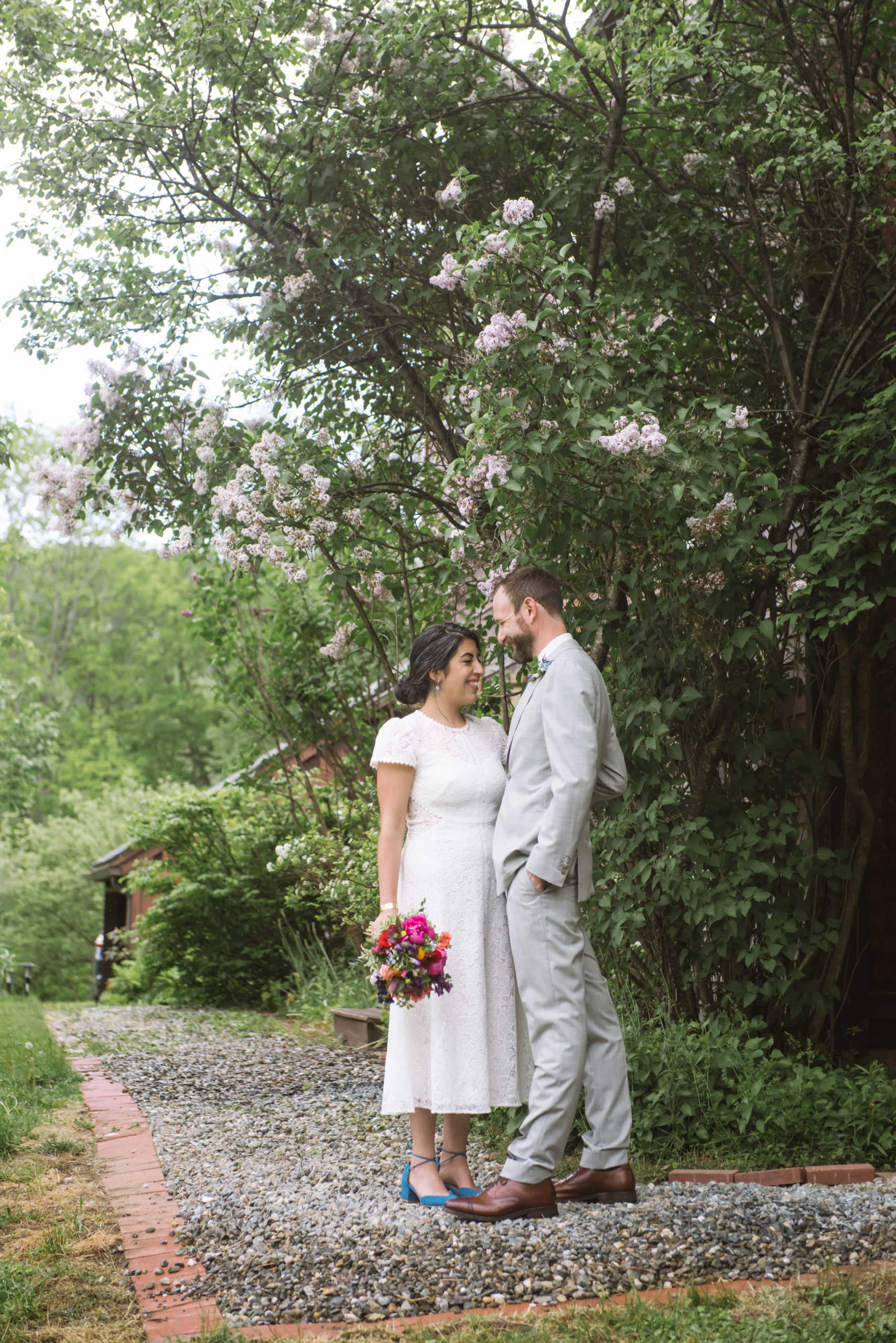 Bride and groom standing facing one another. Both have their eyes closed in happiness and are smiling. The bride is holding her multi-colored bouquet in her right hand and the groom's left hand is in his pocket. They are standing on a gravel pathway in front of flowering greenery.
