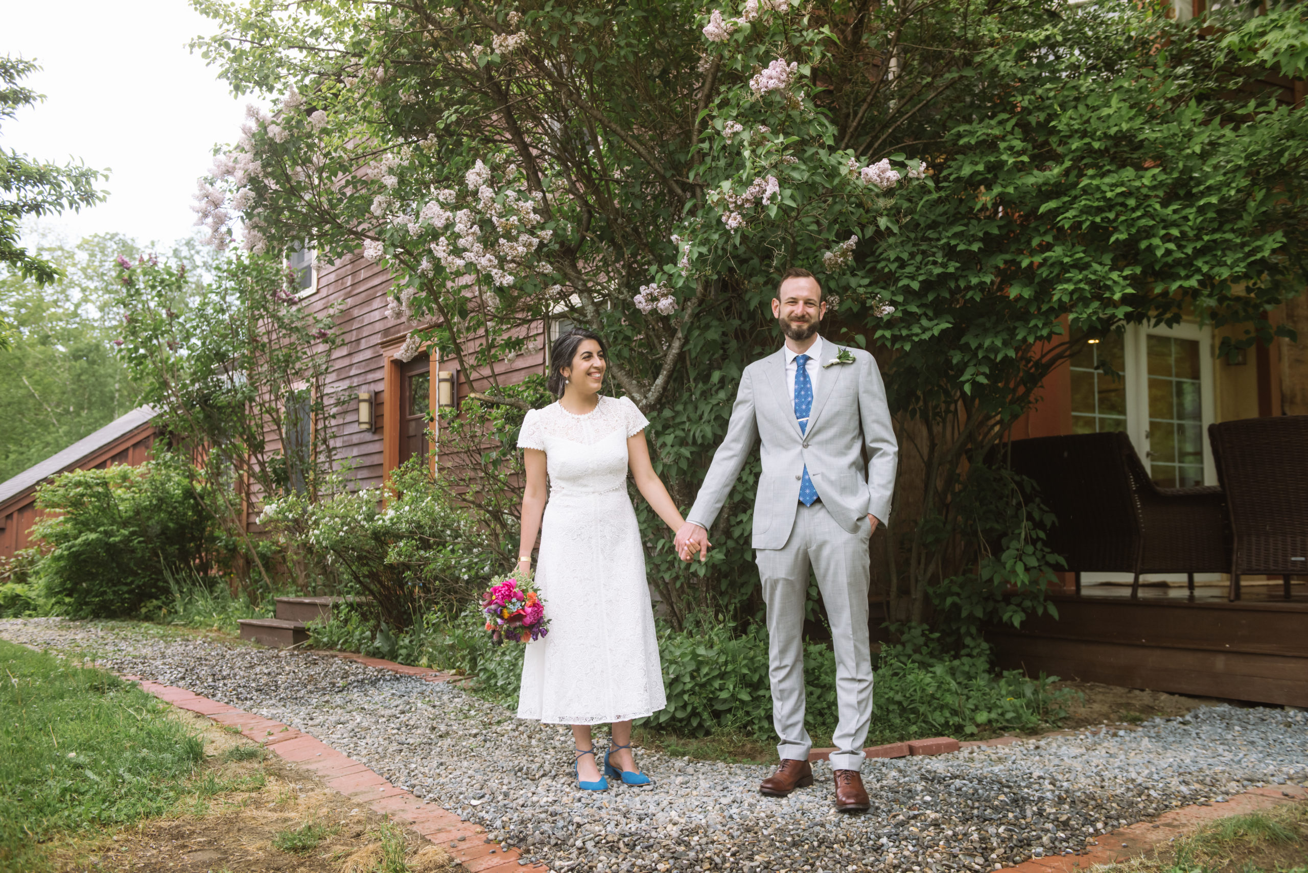 Bride and groom standing apart, holding hands while one is smiling and looking directly to camera and the other is smiling and looking to her groom. Bride is in a white dress and blue shoes, holding a multi-colored pink/purple bouquet. The groom is wearing a light grey suit, blue patterned tie, gold tie clip, white collared shirt, and a boutonniere. They are standing in front of a dark red wood house's front door with greenery and trees surrounding the facade.