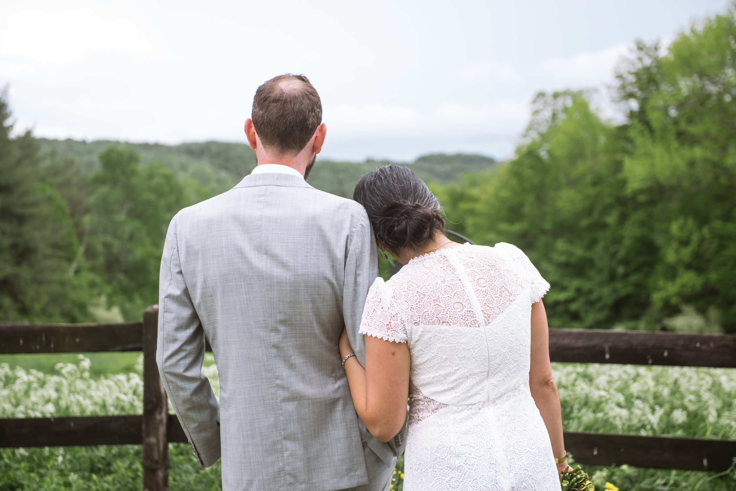 Bride and groom standing side by side with her left arm linked in his right. They are facing away from the camera, facing the mountains, trees, and flowering field in front of them. Her head is rested lightly on his right shoulder. Her bouquet stems are visible from her holding it loosely at her right side.