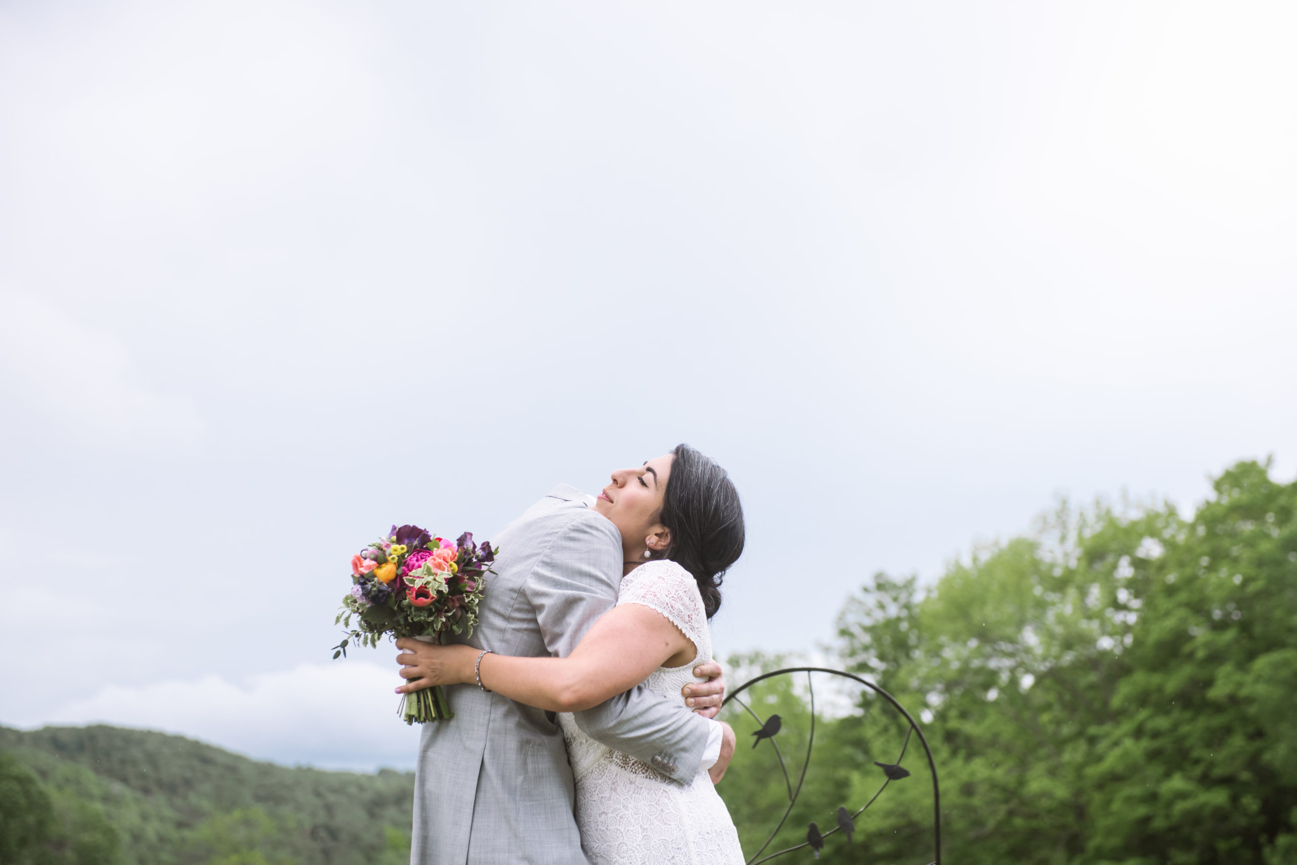 Bride and groom standing, embracing, facing one another. They are kissing one another and have their arms wrapped around each other. The bride is holding her bouquet in her left hand. They are standing in front of a dark wooden fence with lots of white wildflowers in the field. There are trees and mountains in the background.
