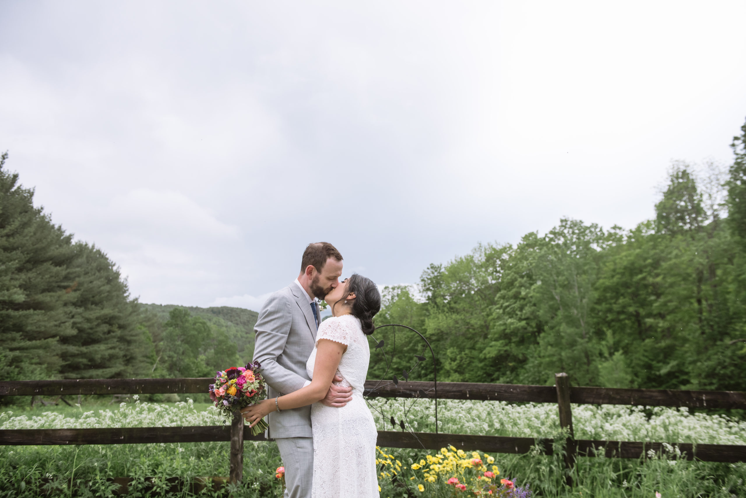 Bride and groom standing, embracing, facing one another. They are kissing one another and have their arms wrapped around each other. The bride is holding her bouquet in her left hand. They are standing in front of a dark wooden fence with lots of white wildflowers in the field. There are trees and mountains in the background.