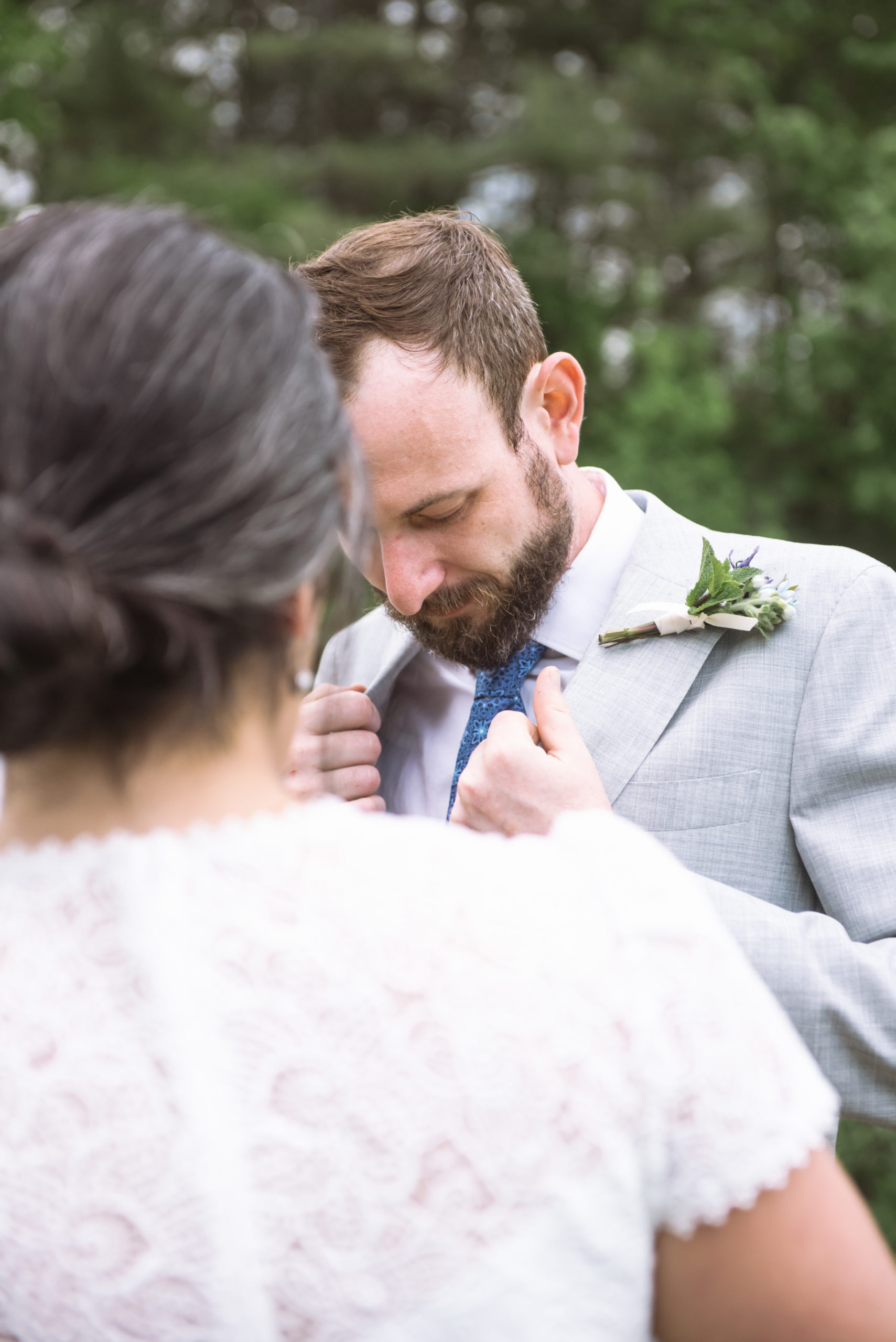 Close up view of the groom looking down, adjusting both sides of the lapels of his suit jacket. He is wearing a blue patterned tie and a boutonniere. The groom is visible behind the bride in the foreground. Her back is to the camera and her hair is in an updo.