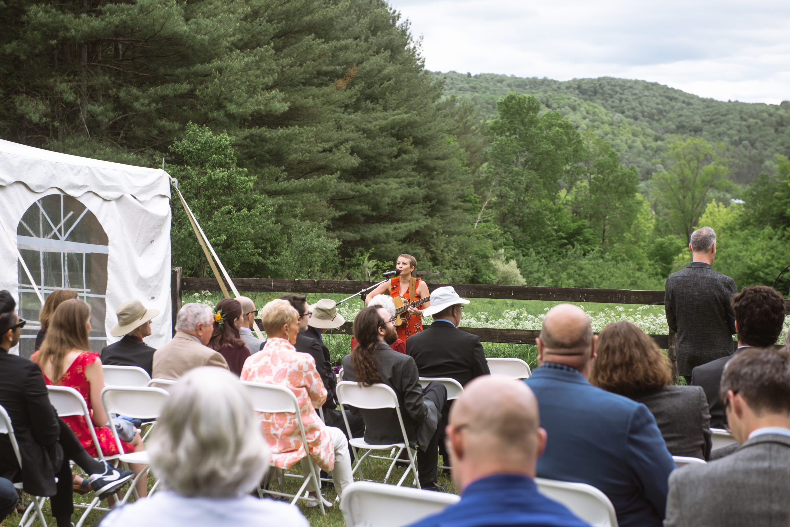 Friend of the couple singing and playing guitar during the ceremony. She is standing in front of a dark wooden fence and there are seated guests in the foreground.