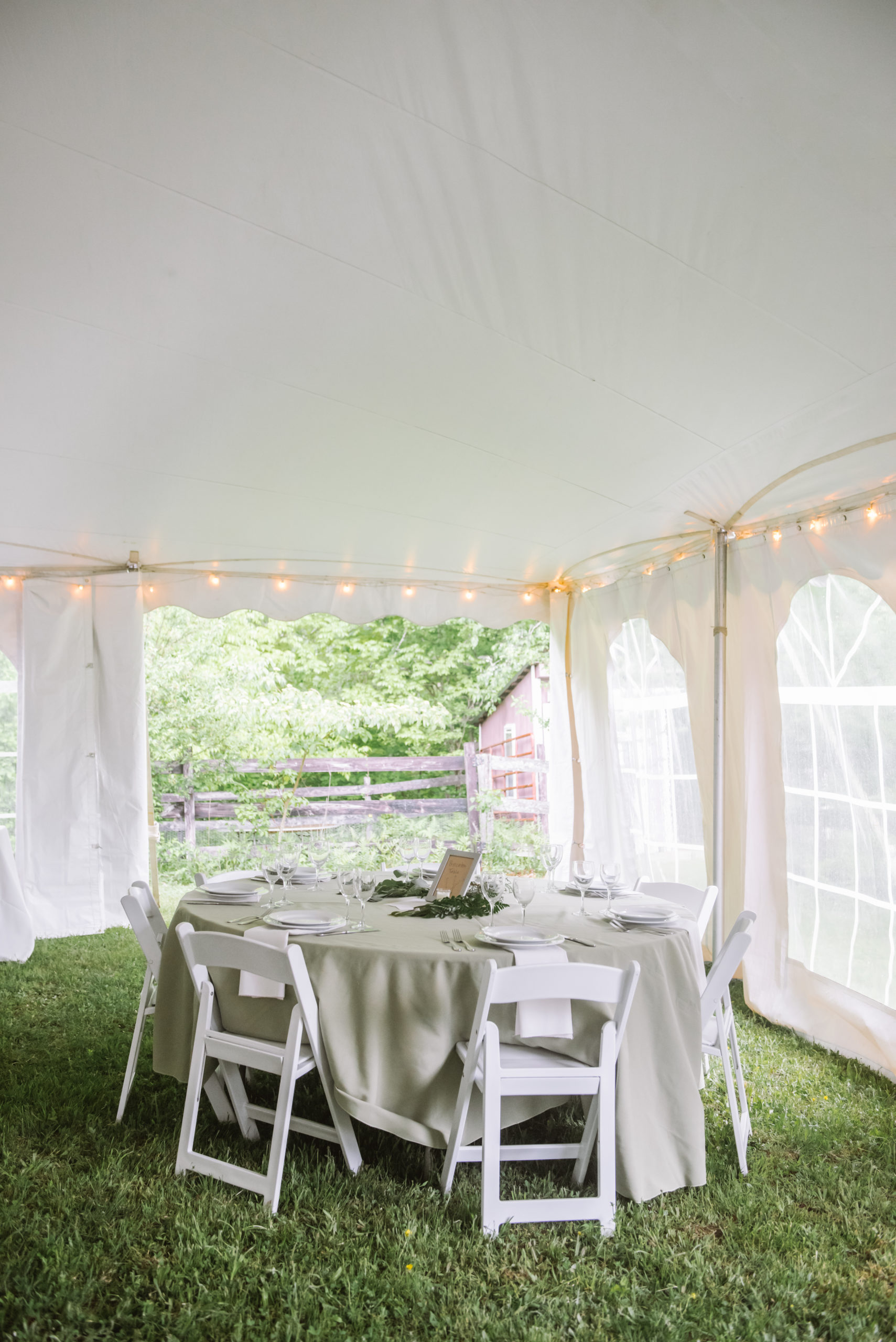 Fully set table in a white reception tent. There is a sage tablecloth, white napkins, and white chairs. The centerpiece is a silver framed brown sign with a wreath of greenery surrounding it.