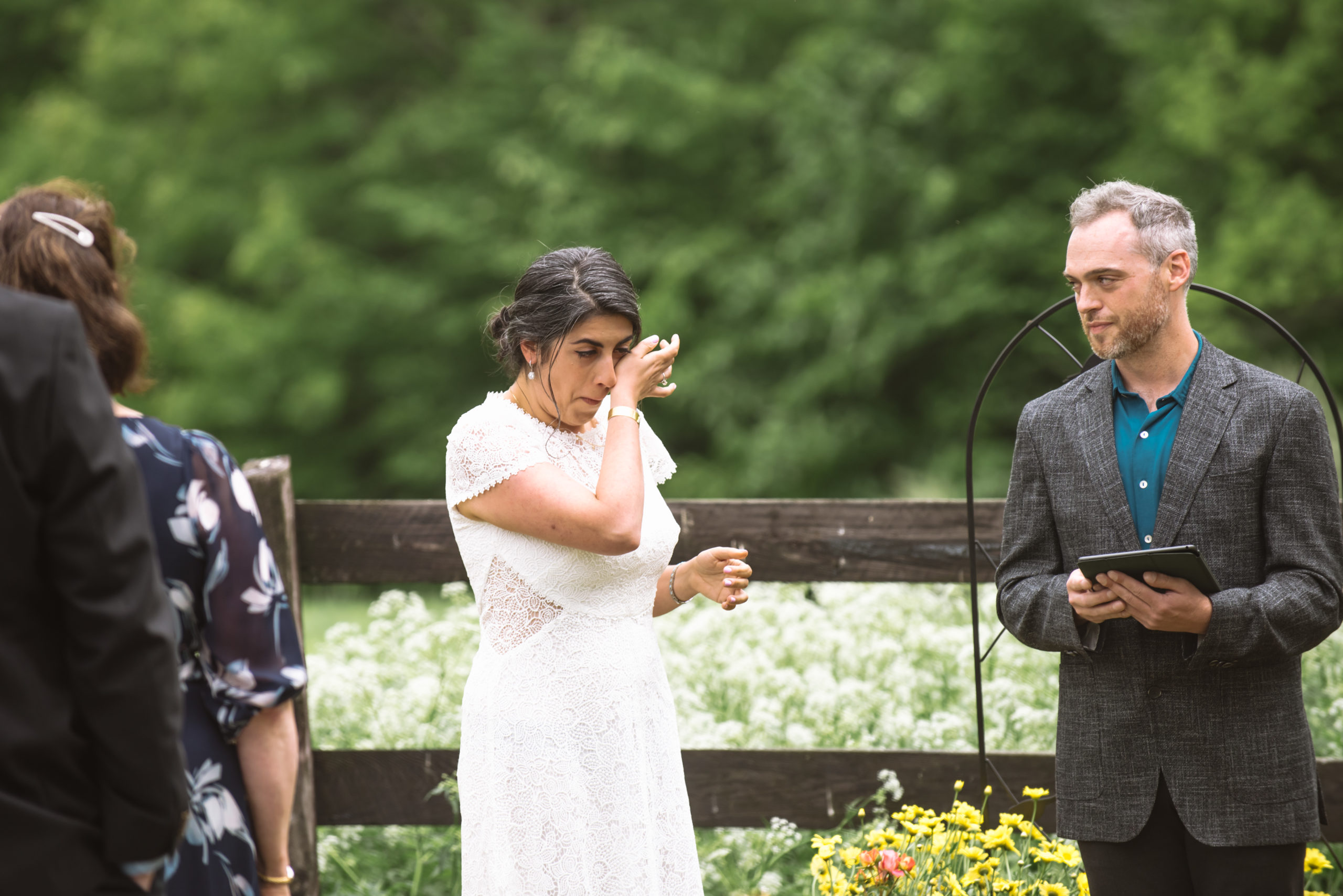 Bride wiping away tears during the ceremony. The officiant is holding an electronic tablet and is looking over at the bride. They are standing in front of a dark wooden fence with lots of white wildflowers in the field. There are trees in the background.