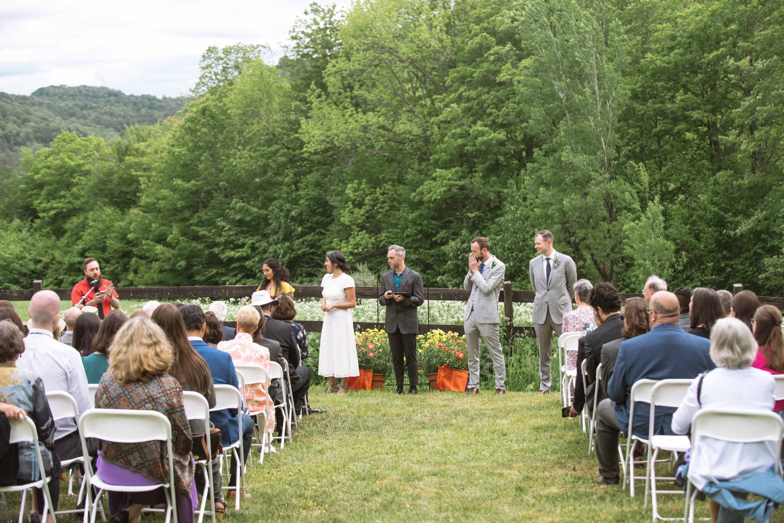 Ceremony featuring the couple and their wedding party (maid of honor and best man) at the alter with seated guests in the foreground. A friend of the couple is reciting a spoken-word poem at the microphone. Everyone is turned to look at him. The groom has his hands in front of his mouth in a moved/emotional manner. They are standing in front of a dark wooden fence with lots of white wildflowers in the field. There are trees and mountains in the background.