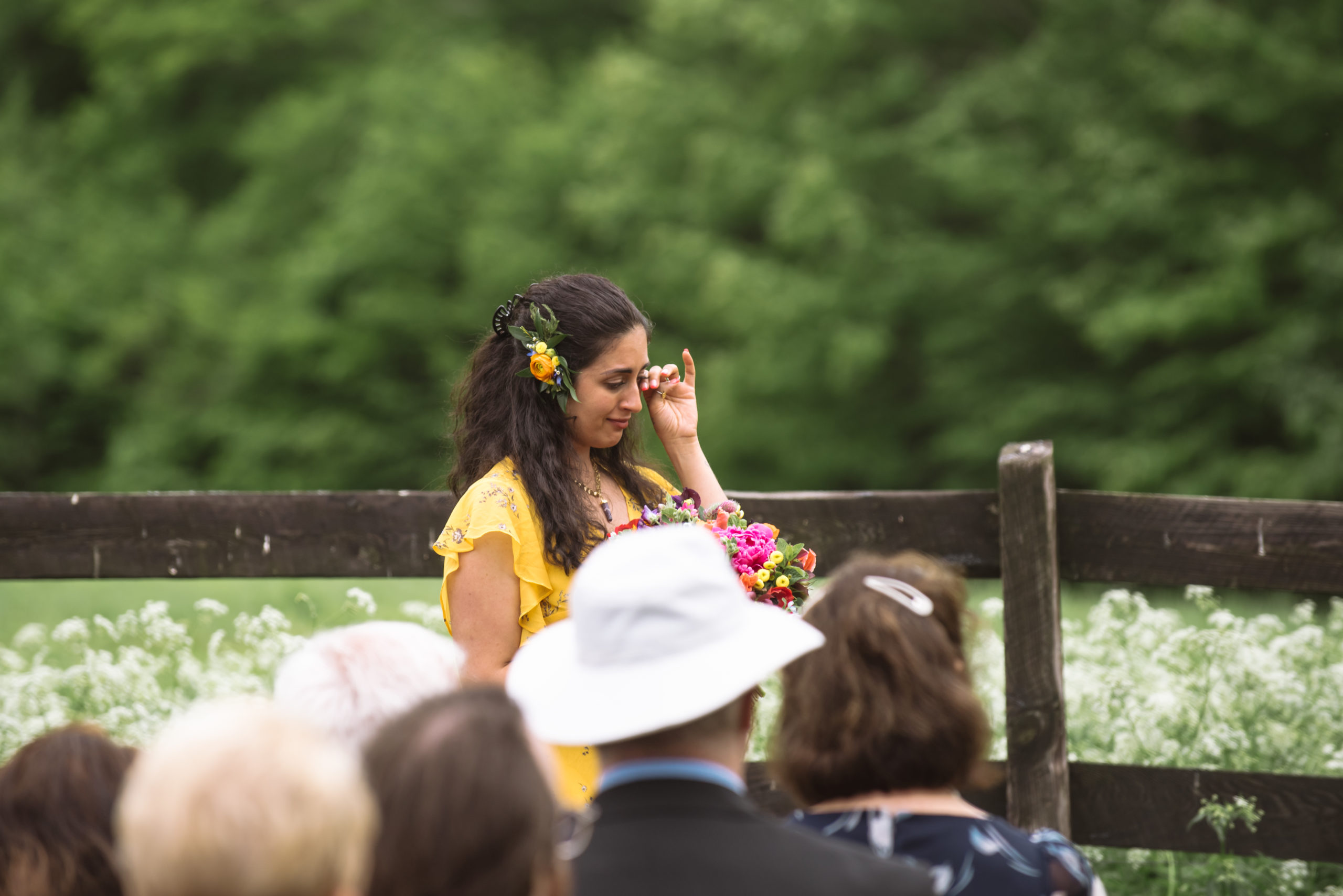 Maid of honor, the bride's sister, wiping away a tear during the ceremony. She is wearing a yellow floral dress with citrus colored florals in her hair. Seated guests are seen in the foreground.