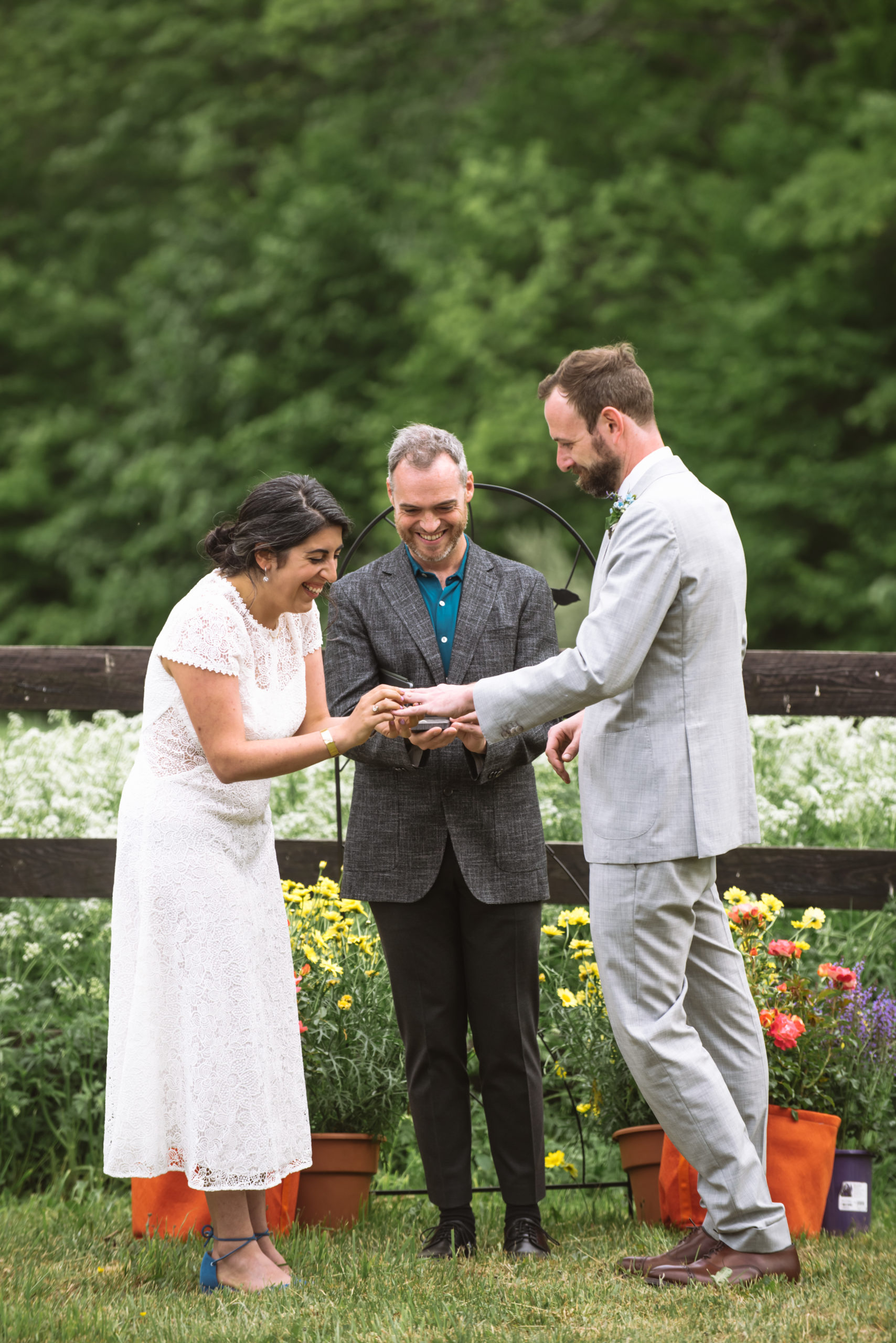 Bride placing the wedding band on her groom. The couple and the officiant are smiling. They are standing in front of a dark wooden fence with lots of white wildflowers in the field. There are trees in the background. 