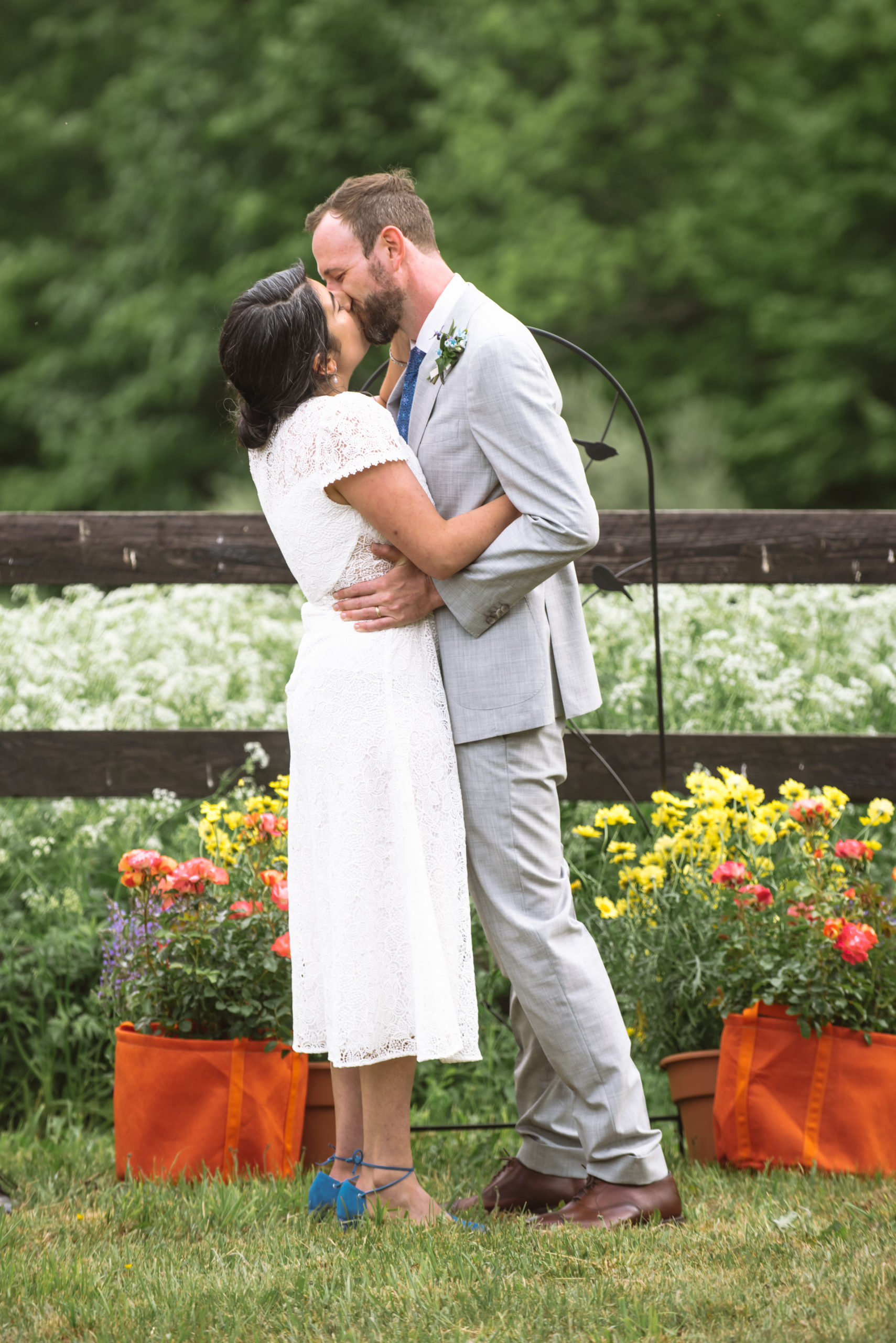 Ceremony featuring the couple embracing with their arms around one another and kissing after being pronounced wife and husband. They are standing in front of a dark wooden fence with lots of white wildflowers in the field. There are trees and mountains in the background.