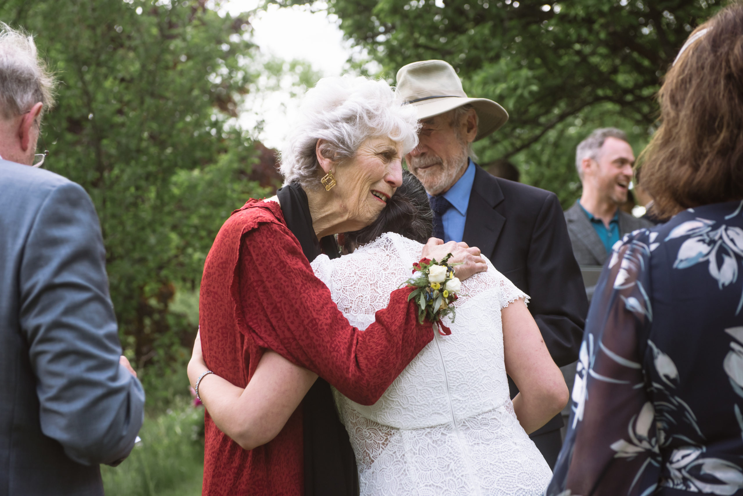 Grandmother of the bride hugging the bride post-ceremony. There are guests surrounding them in the foreground and background. Trees are in the far background.