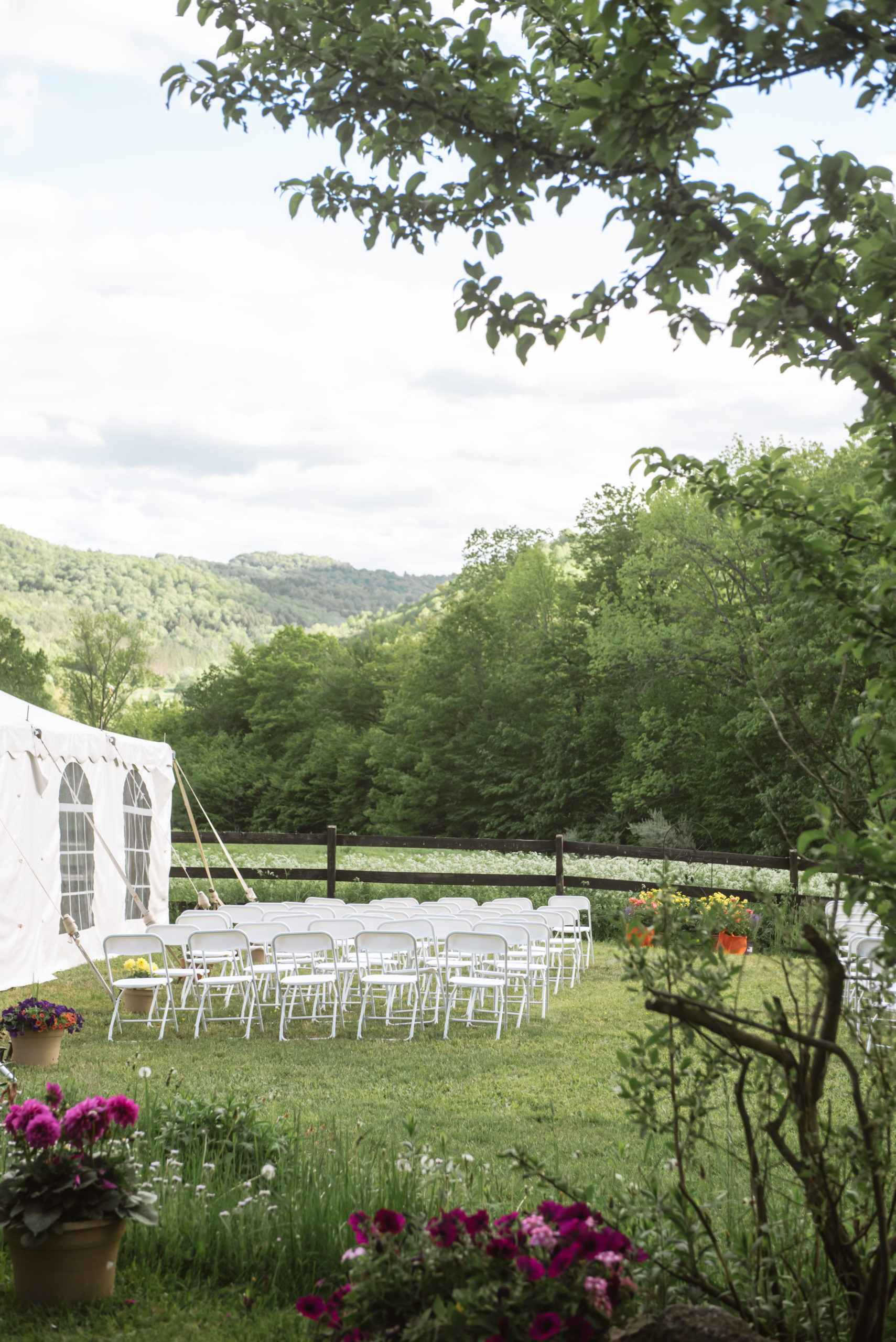 Ceremony seats lined up in a field next to a white tent and in front of a dark wooden fence. There is a white floral field, trees, and mountains in the background.