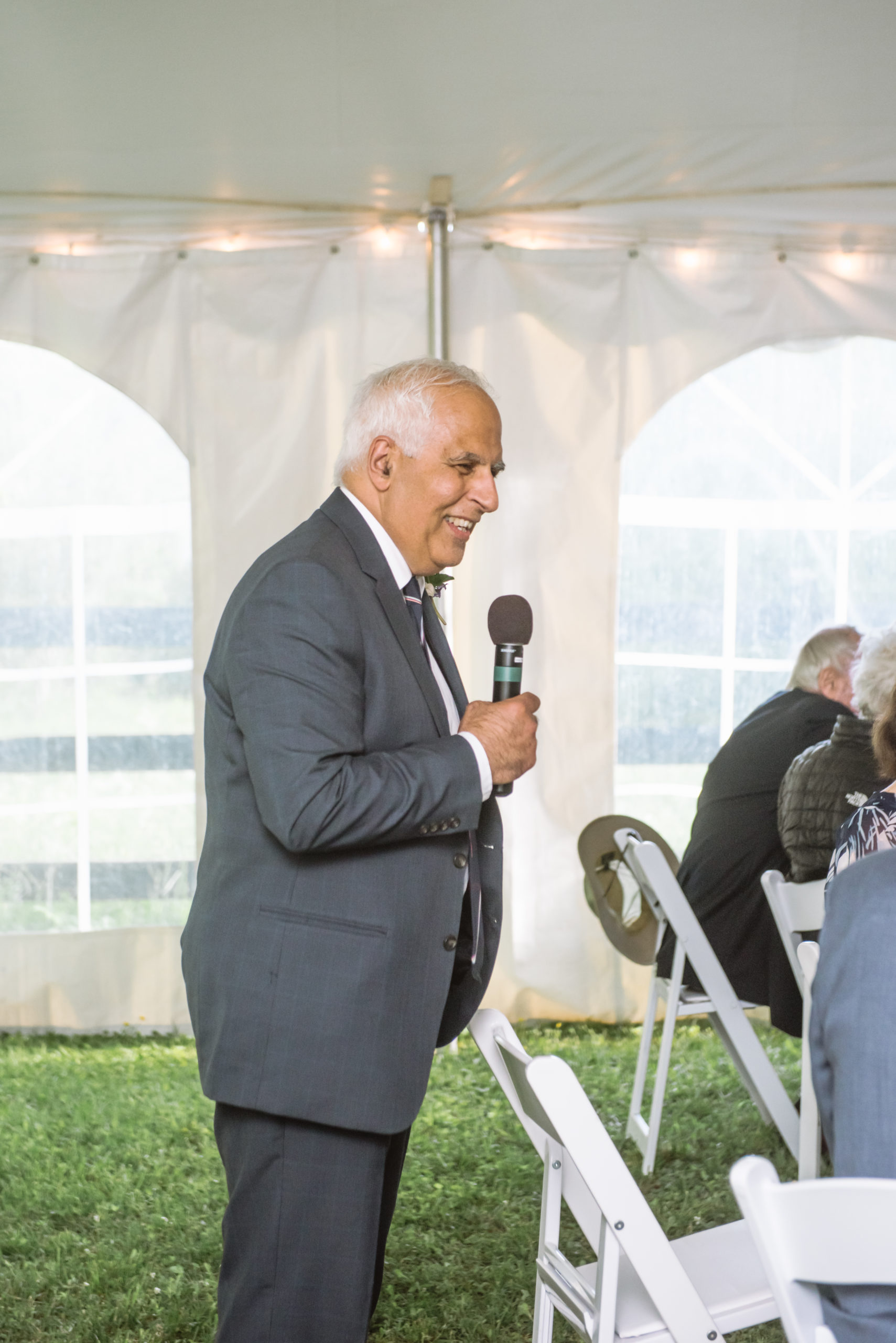 Father of the bride speaking into a microphone for a speech. He has a huge smile on his face and is standing behind a white chair in a white tent.