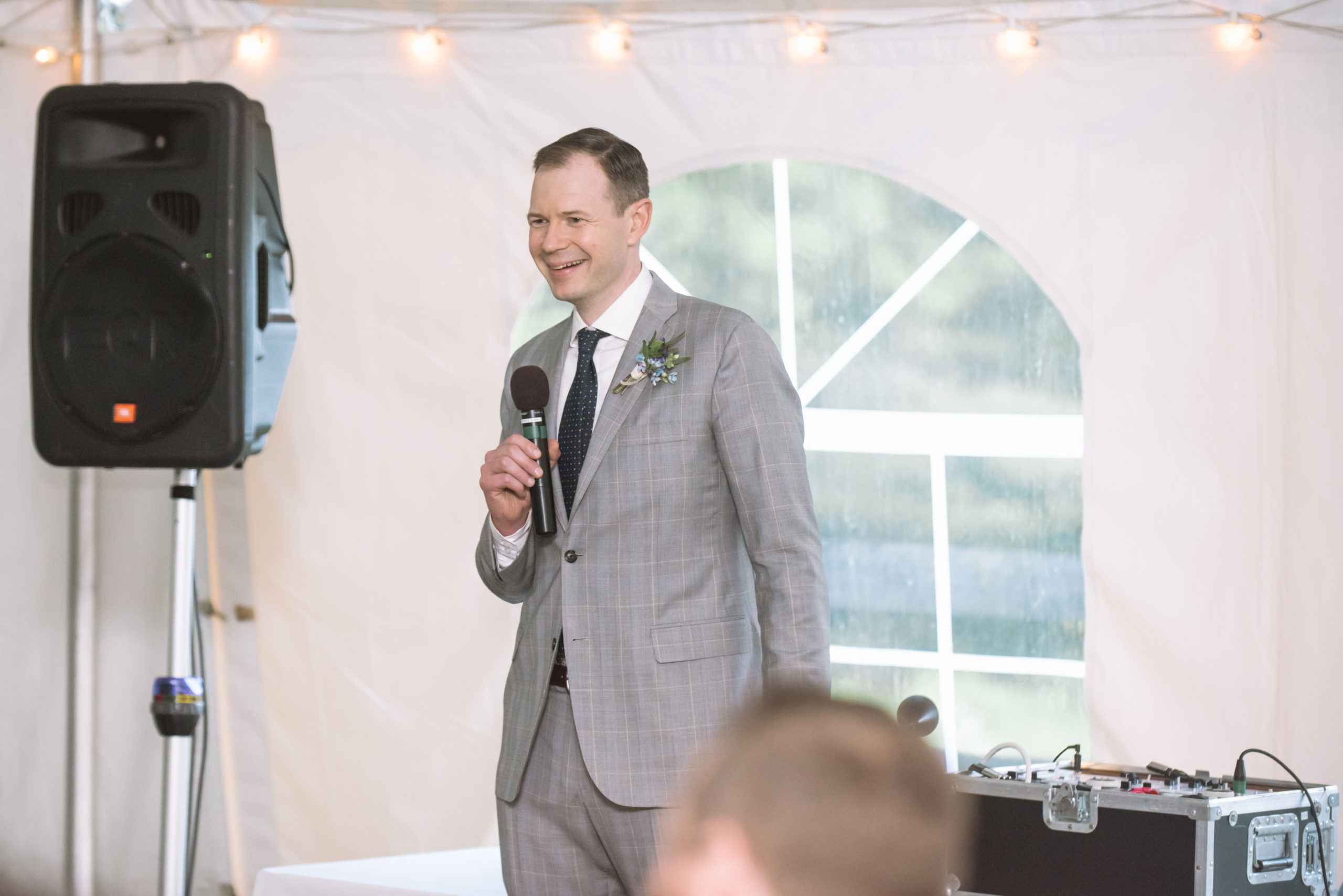 Brother of the groom, the best man, giving a speech during the tented-reception. He is holding a microphone and is open-mouthed smiling.