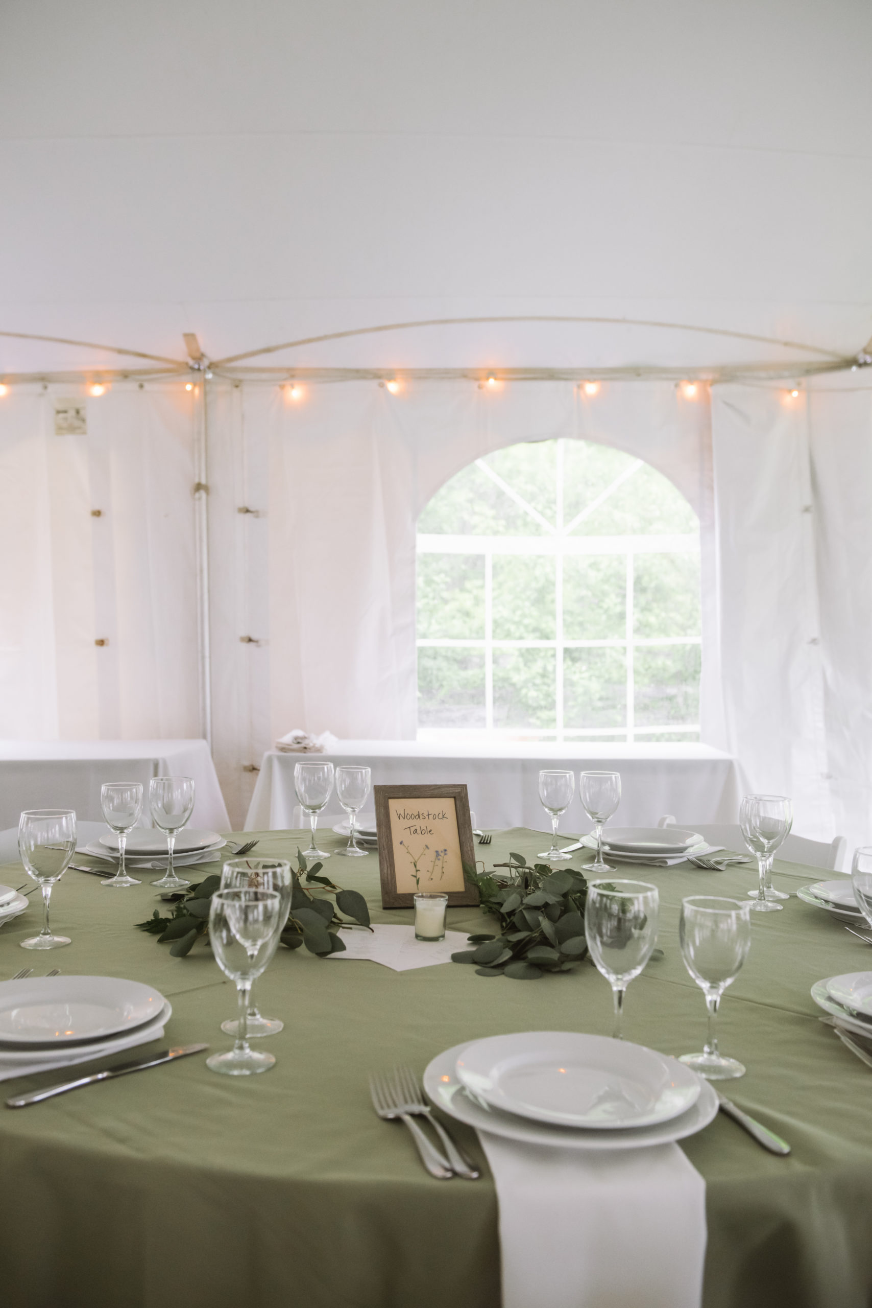 Semi-close view of the "Woodstock Table" at the tented wedding reception. The table is fully set with plates, silverware, glasses, and napkins. There is a silver framed sign with a wreath of greenery around it.