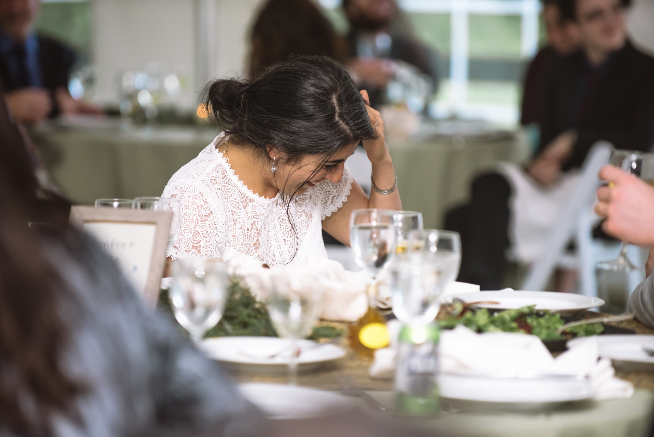 Bride hearing a speech during the white tent reception. She is laughing while looking down toward her lap and touching her hair. There are guests all around in the foreground and background.