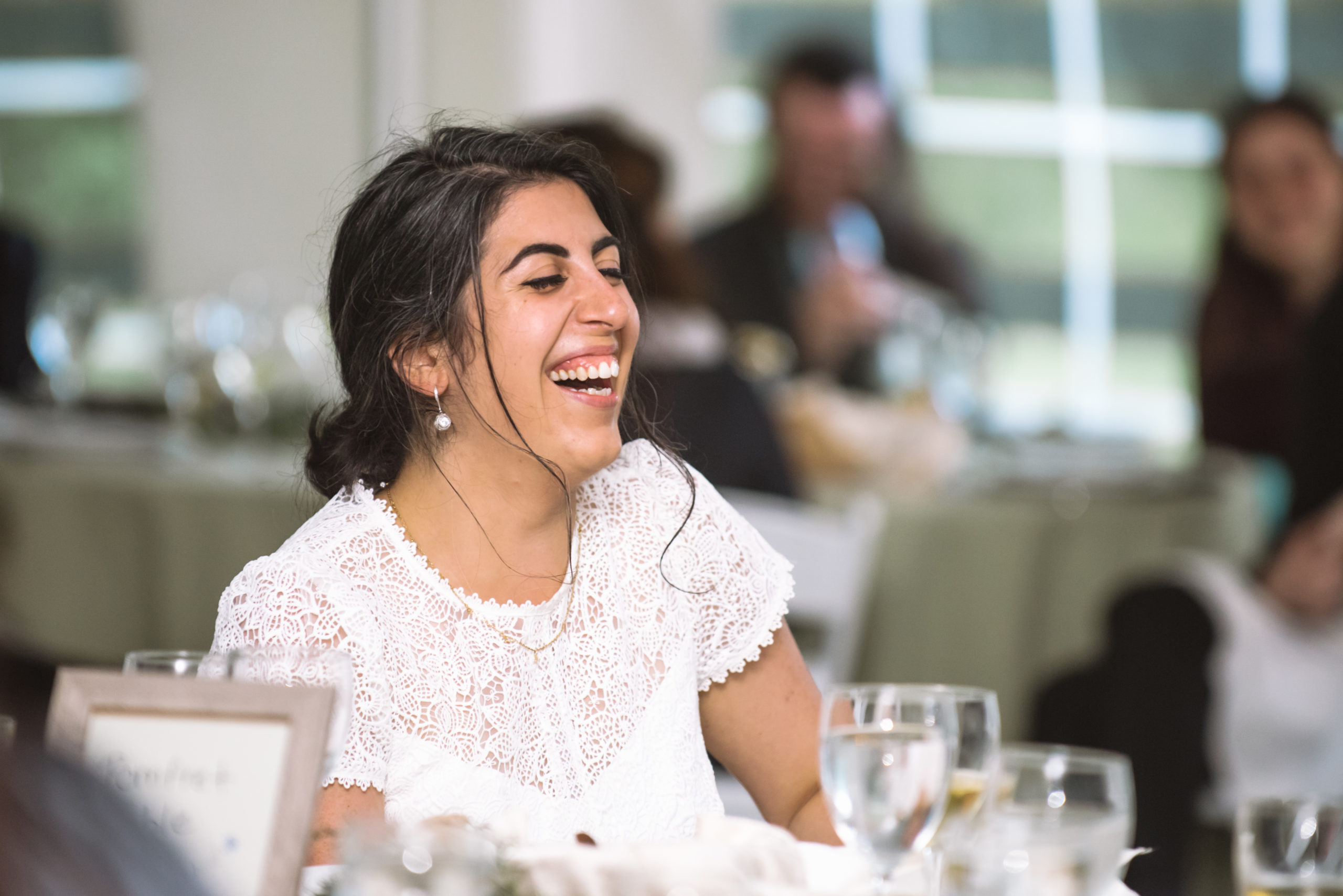 Bride hearing a speech during the white tent reception. She is open-mouthed laughing with her eyes closed. There are guests all around in the background.