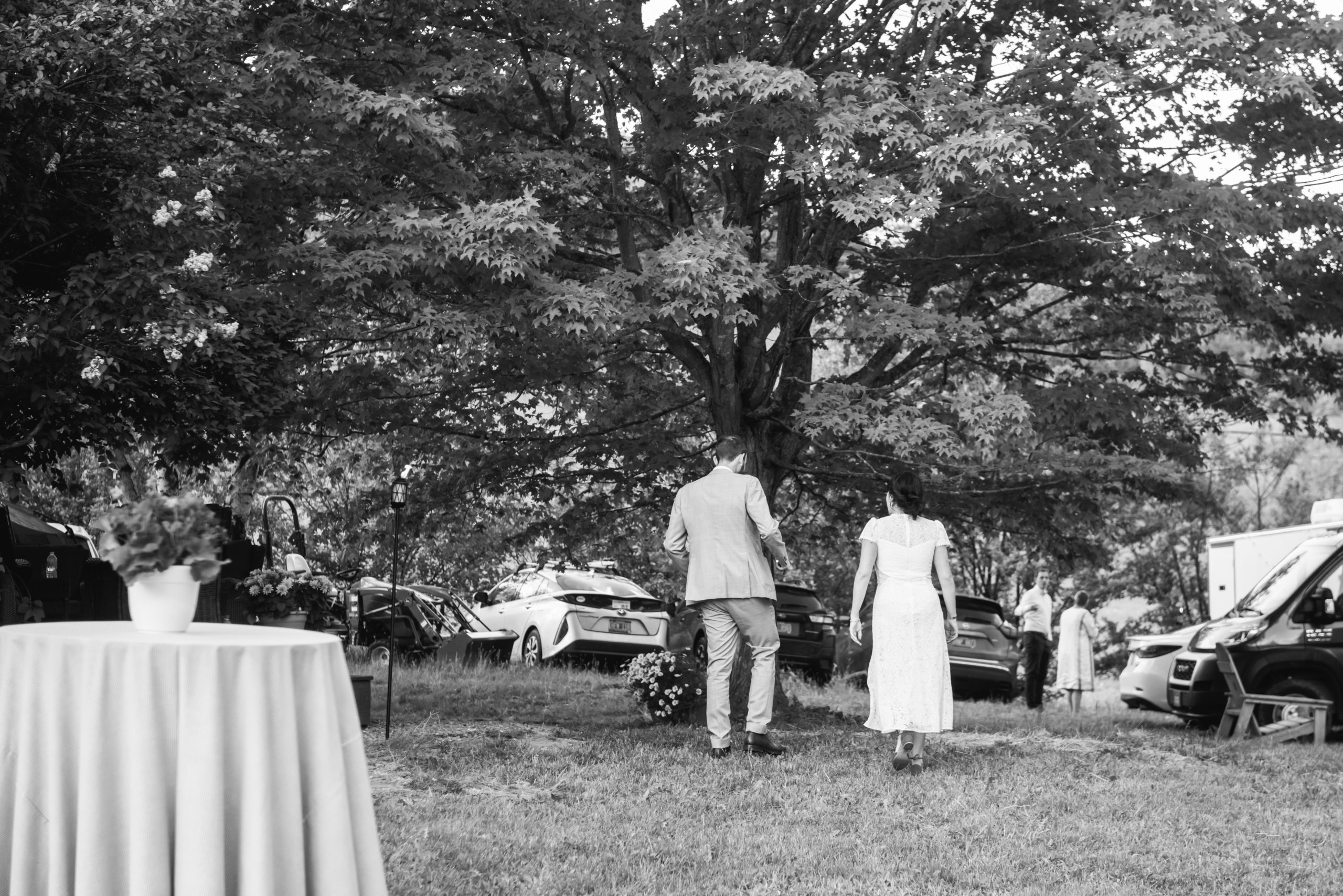 Black and white photo of the bride and groom walking away from the reception tent (not visible in the photo). They are mid-walk in grass with their backs away from the camera. There is a big tree in front of them and there are cars parked in the background.
