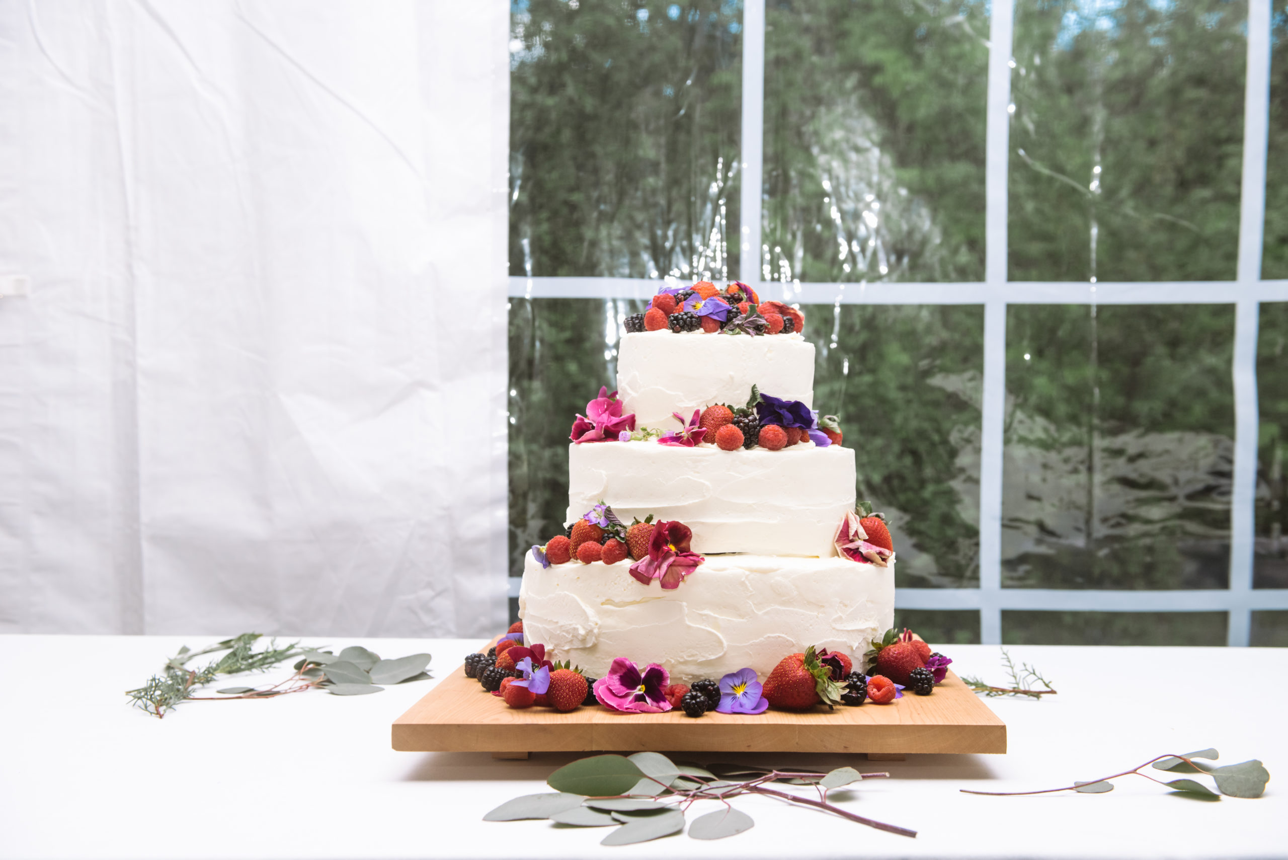 Three-tiered white cake adorned with fresh raspberries, strawberries, blackberries, and pink and purple flowers. The cake is on a square wooden cake stand and there is some greenery around the cake. It is set on a white tablecloth and it is within a white tent.