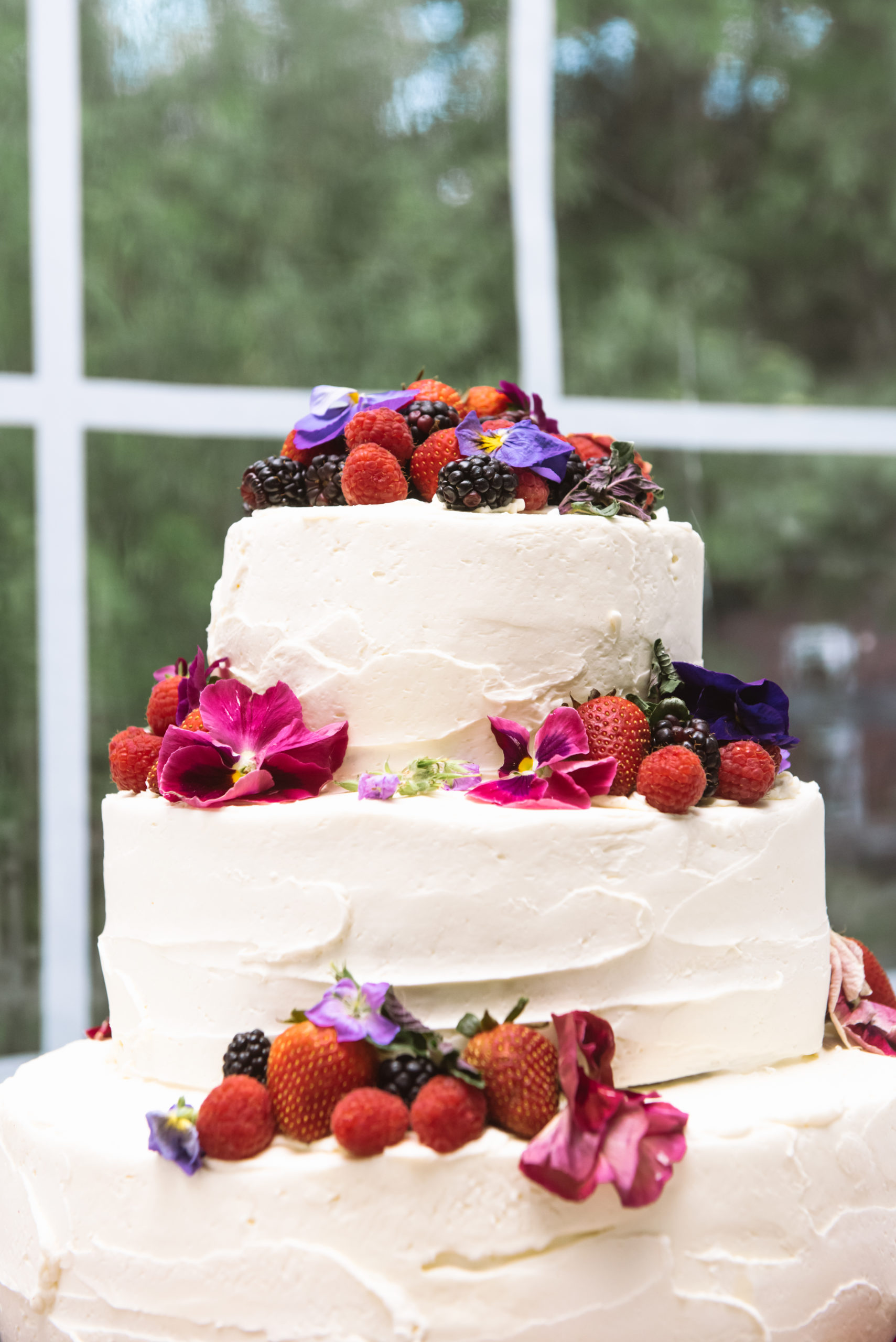 Close up on the top two tiers of a three-tiered white cake adorned with fresh raspberries, strawberries, blackberries, and pink and purple flowers. It is within a white tent.