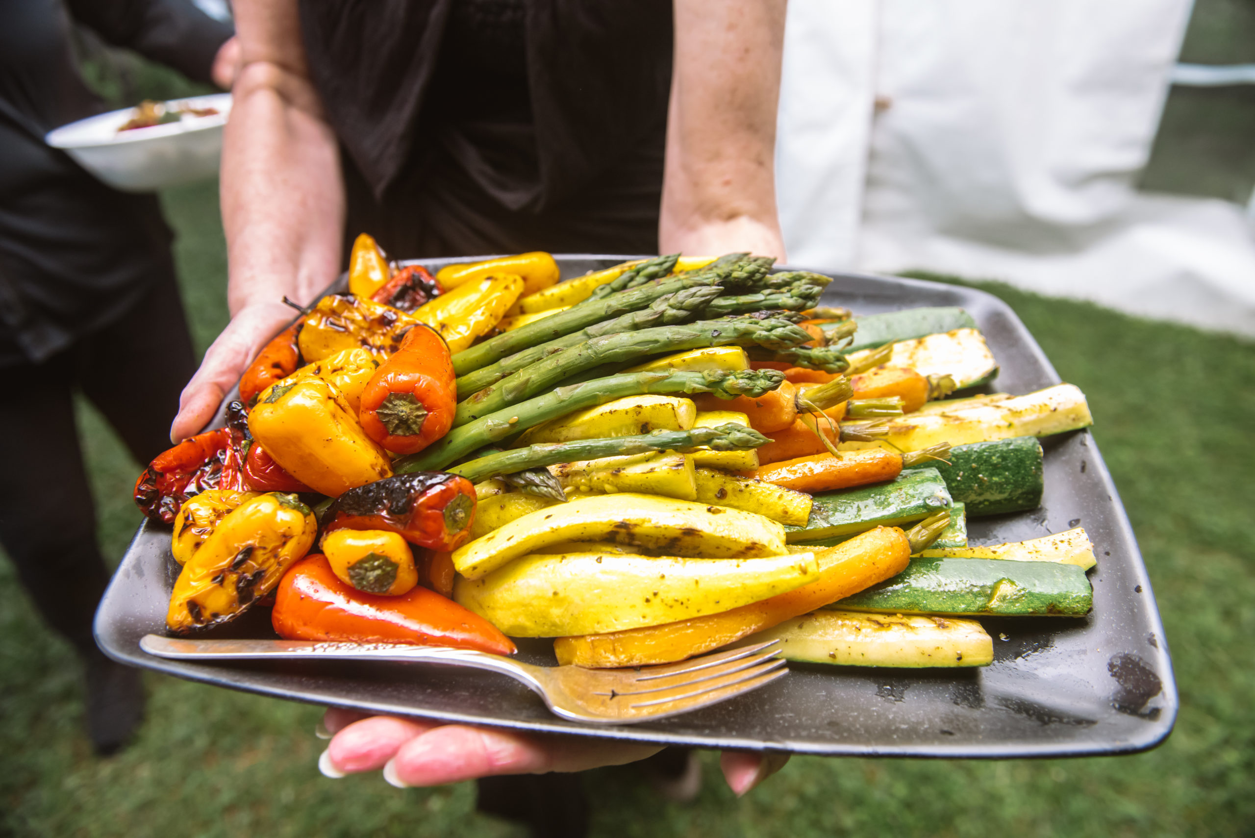 Server holding up a black family-style serving tray with lots of roasted vegetables including peppers, asparagus, zucchini, carrots, and squash. There is a silver fork laid on the tray.
