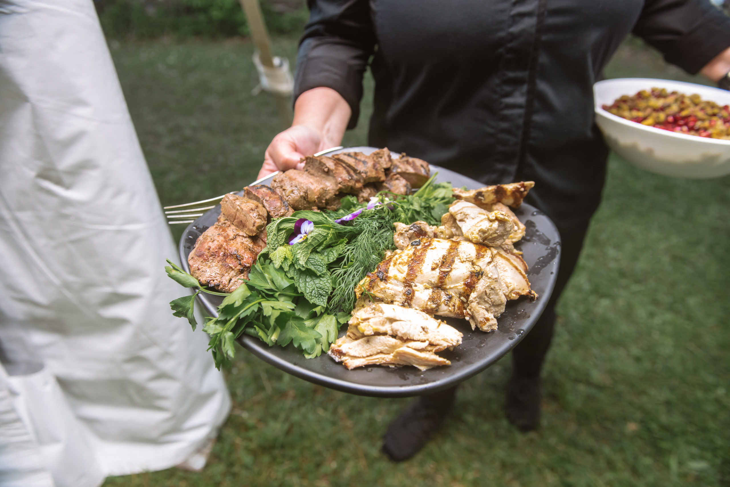 Close up view of a server holding a black serving platter in their right hand. It has grilled chicken breasts, herbs, and a darker meat. In the server's left hand is of the white family-style serving bowls in their left hand featuring pomegranate seeds and green/brown seeds. 