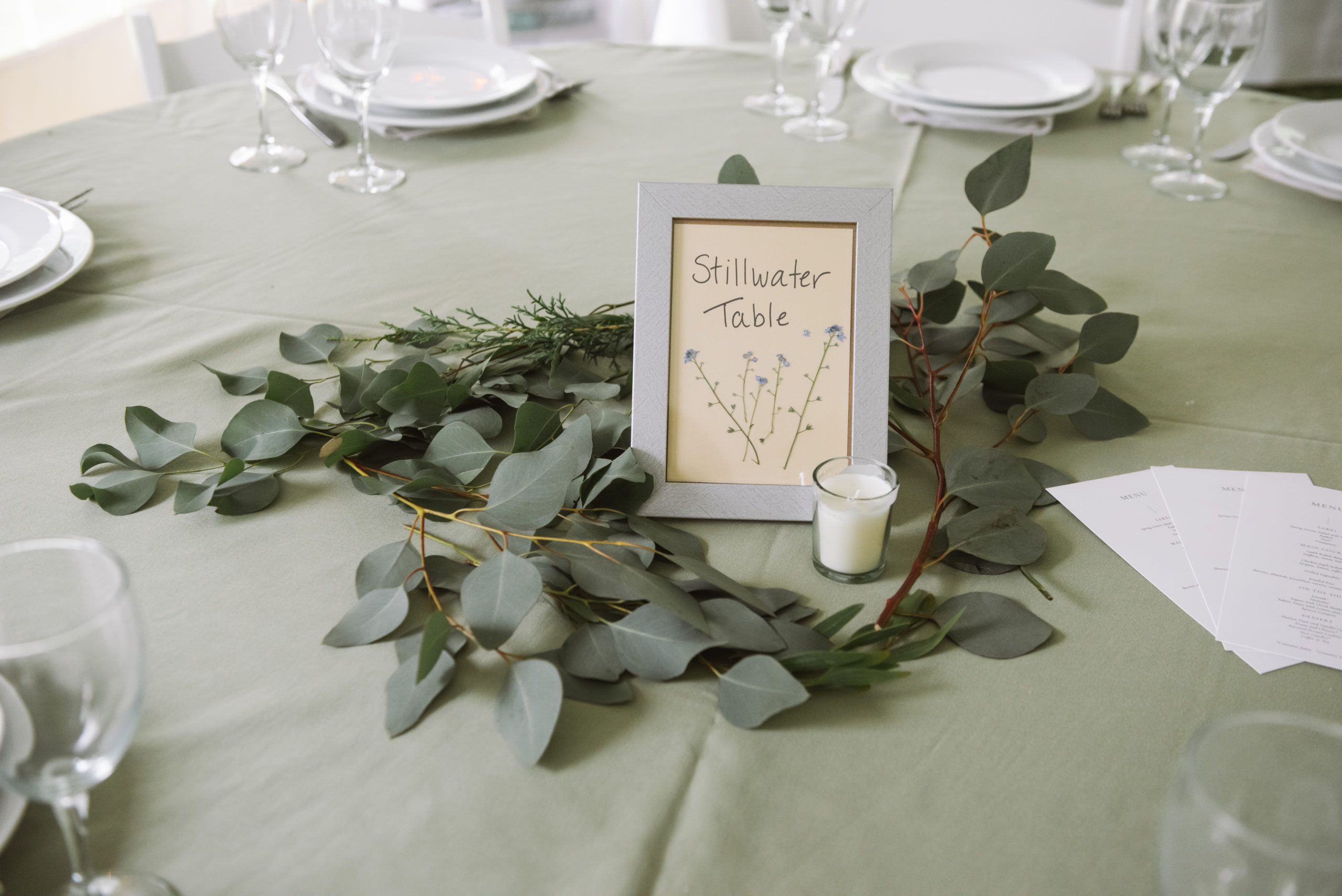 "Stillwater Table" silver framed table sign with dried flowers under the name. The frame is surrounded by greenery and an unlit white small candle. It is in the center of a reception table.