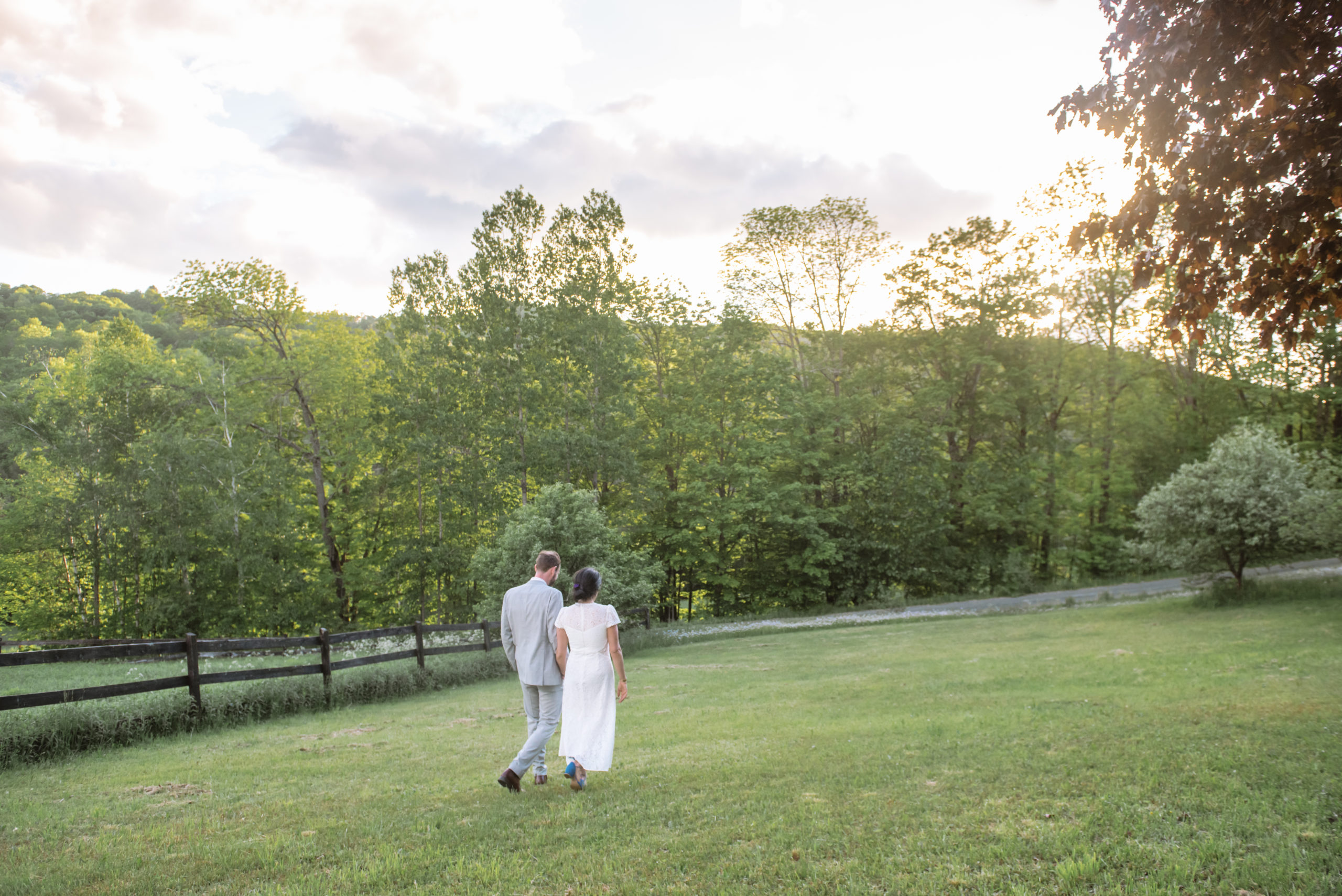 Bride and groom walking away from the camera holding hands. They are walking in a field in front of a dark wooden fence encasing white flowers. The setting sun is shining light through the trees and over the mountain in the background.