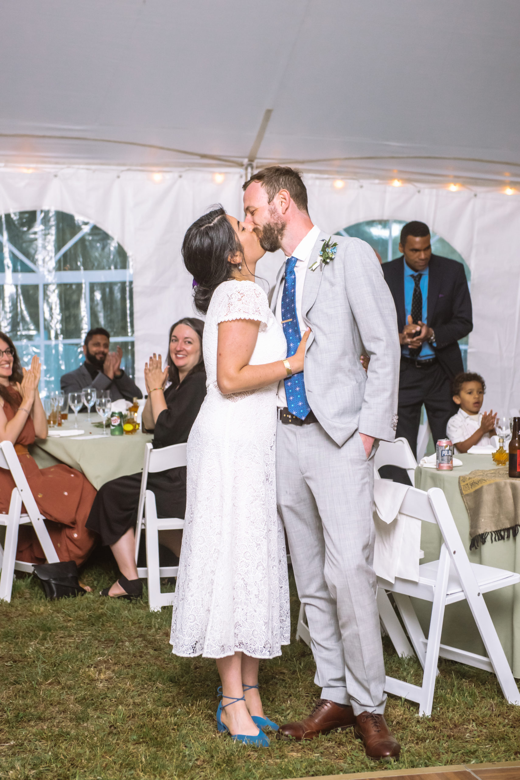 Bride and groom kissing during their reception. They are embracing face to face with the arms around each other. Both have their eyes closed. There are wedding guests clapping in the background.
