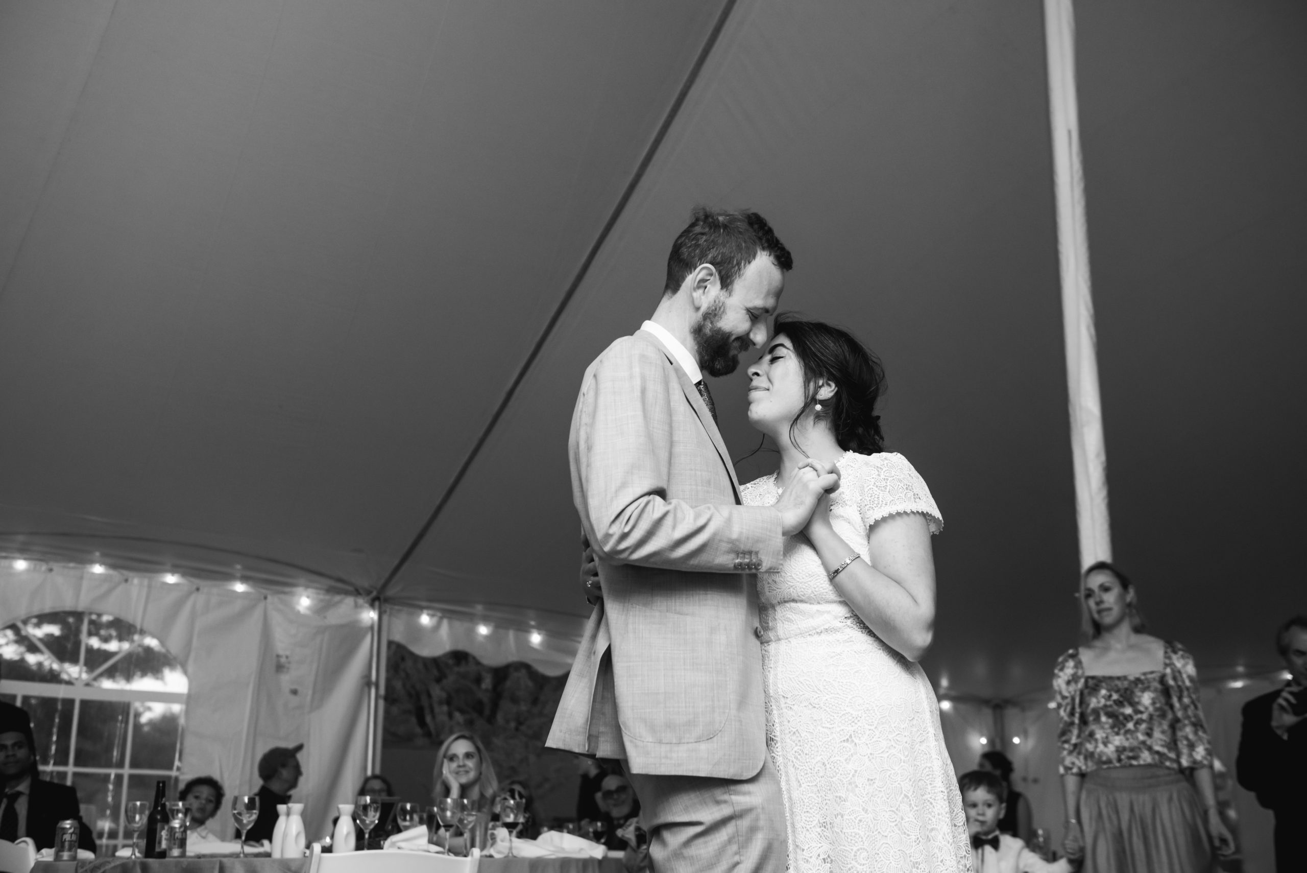 Black and white photo of the couple slow dancing during their reception. They are embracing face to face with the arms around each other and holding hands. Both have their eyes closed and have soft smiles in a tender moment. There are wedding guests in the background.