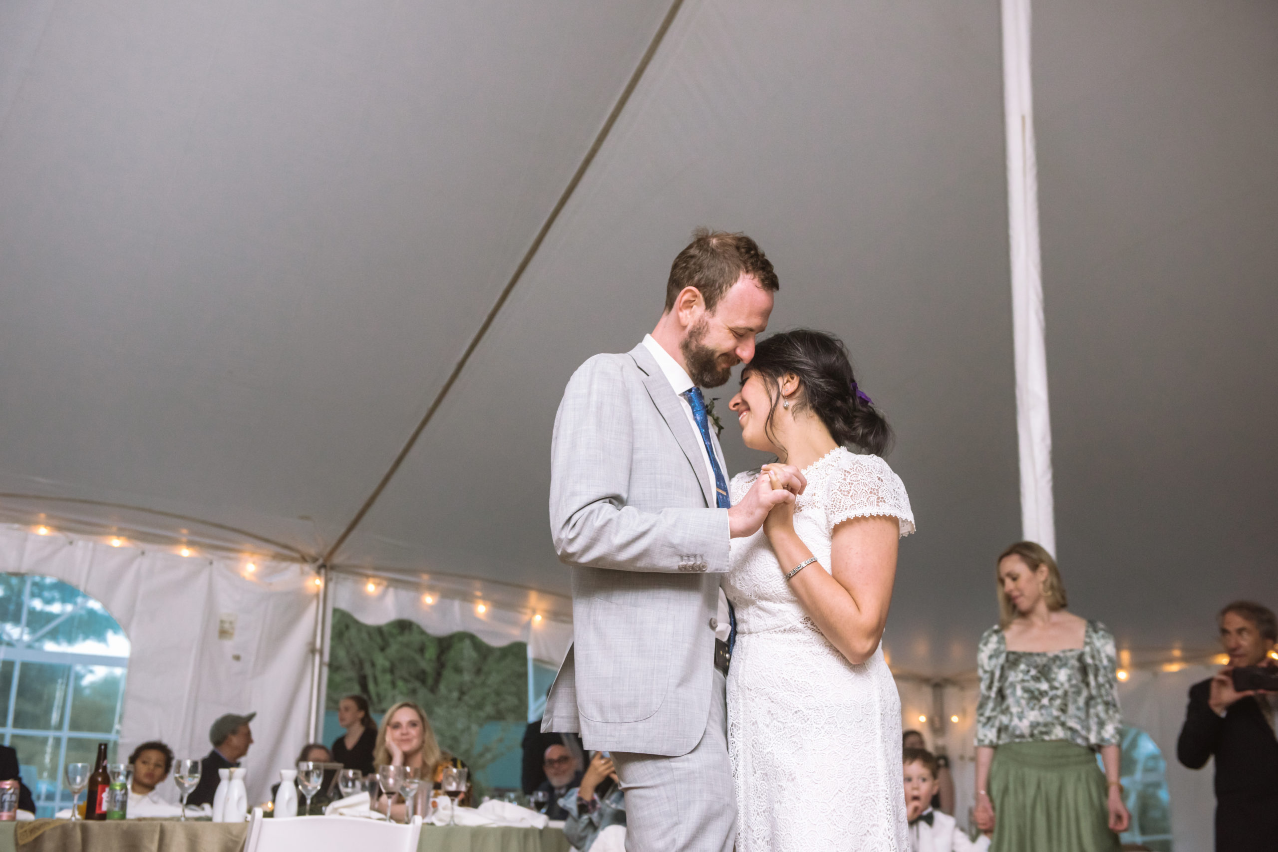The newly married couple slow dancing during their reception. They are embracing face to face with the arms around each other and holding hands. Both have their eyes closed and are smiling in a tender moment. There are wedding guests in the background.