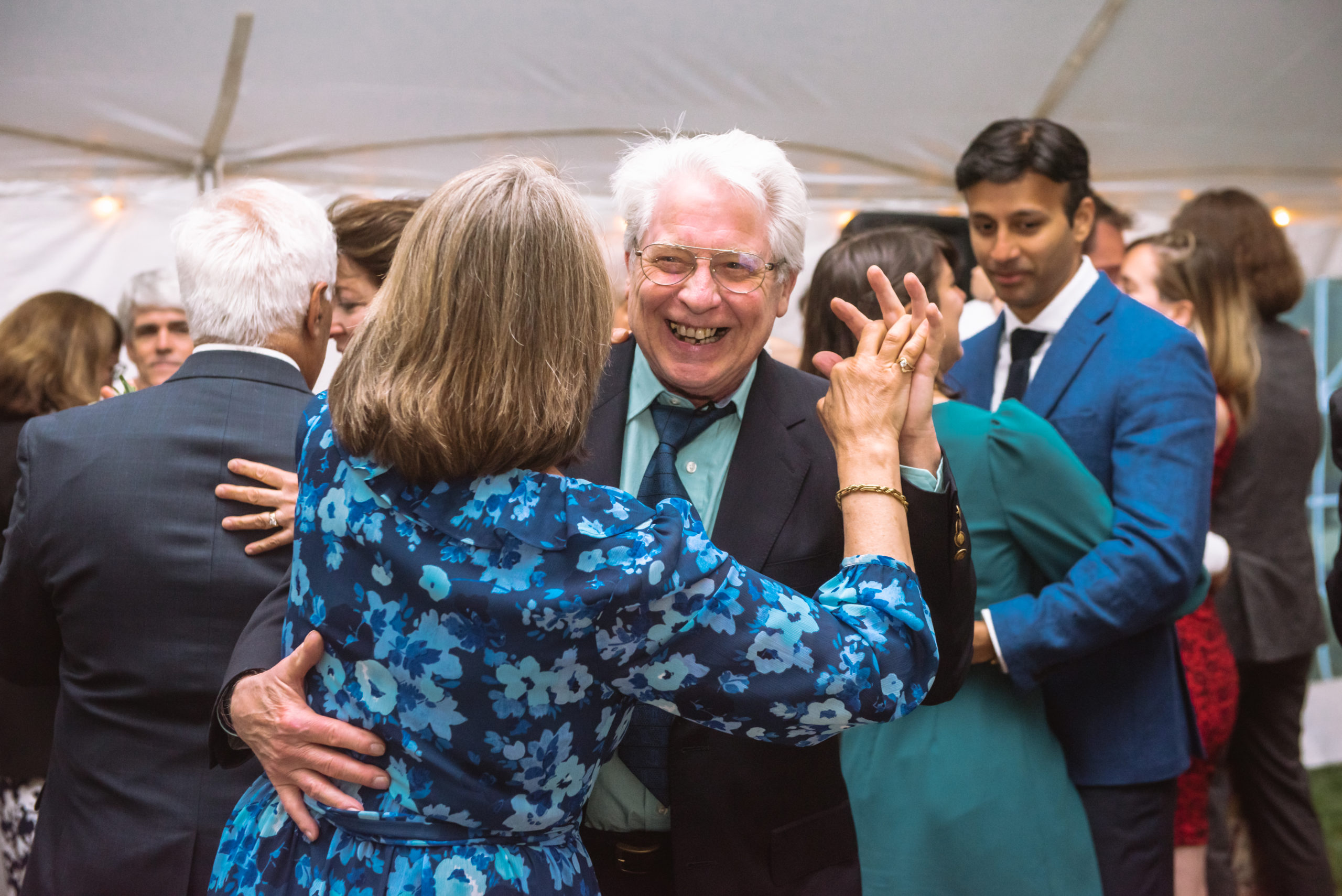 Wedding guests slow-dancing in the tented reception. There is an older couple front and center. The woman's back is turned away from the camera and her partner is open-mouthed grinning.