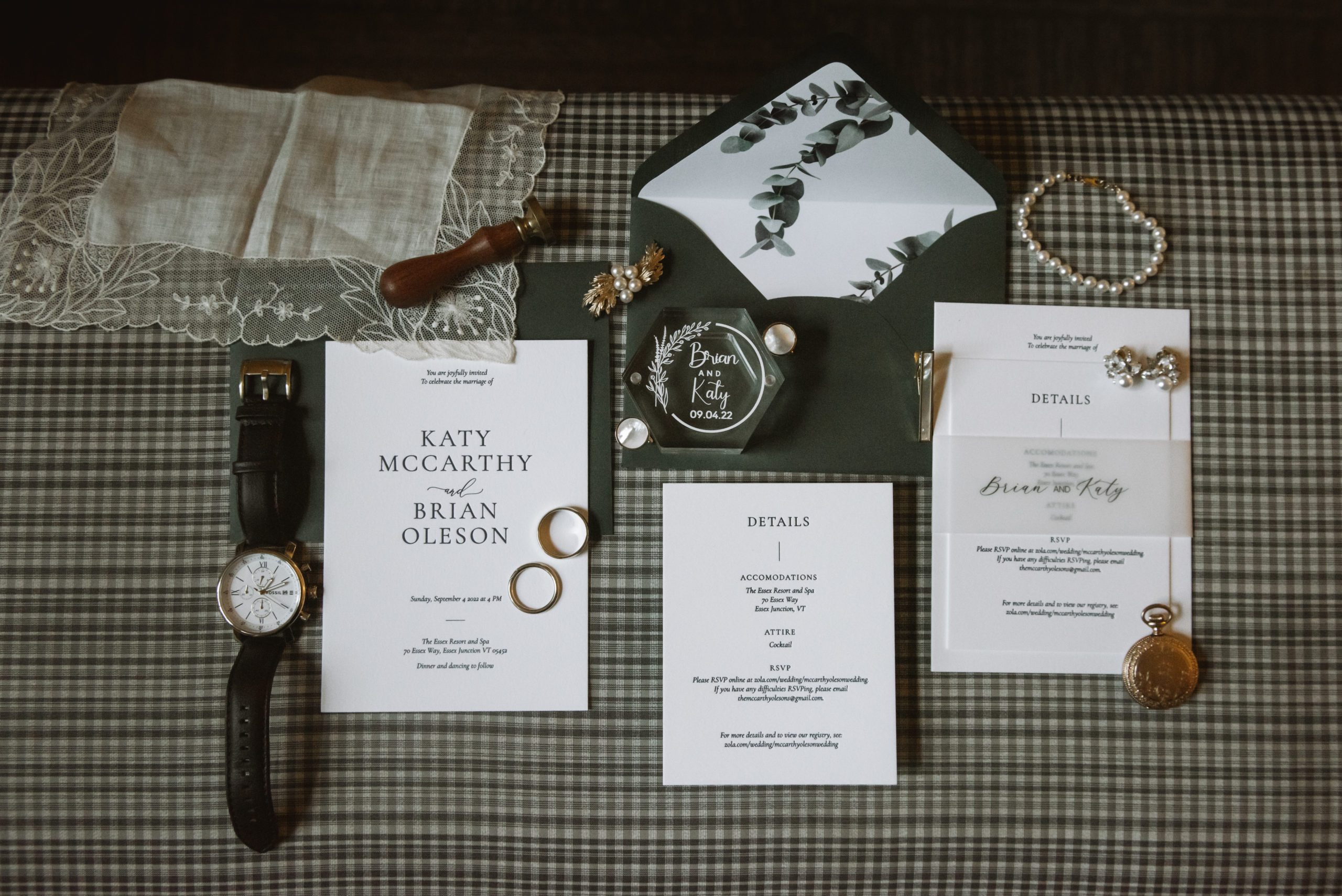 Lay flat of the couple's dark green stationery. Included is the invitation, detail card, envelopes, and the bride and groom's details like jewelry, rings, and lace handkerchief. Everything is set atop a green, black, and white gingham ottoman.