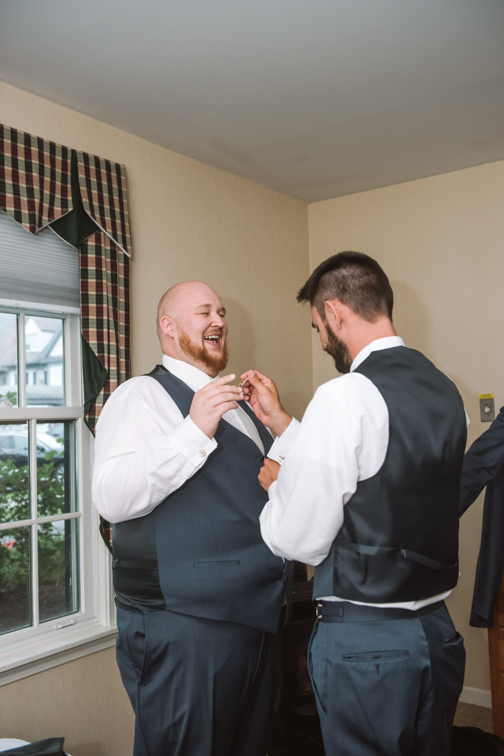 Groom getting help with his tie clip from one of his groomsmen. Theya re standing face to face in a hotel room. The groomsmen is looking down at what he's doing with his hands while the groom is laughing in a big open-mouth smile looking at his groomsman.