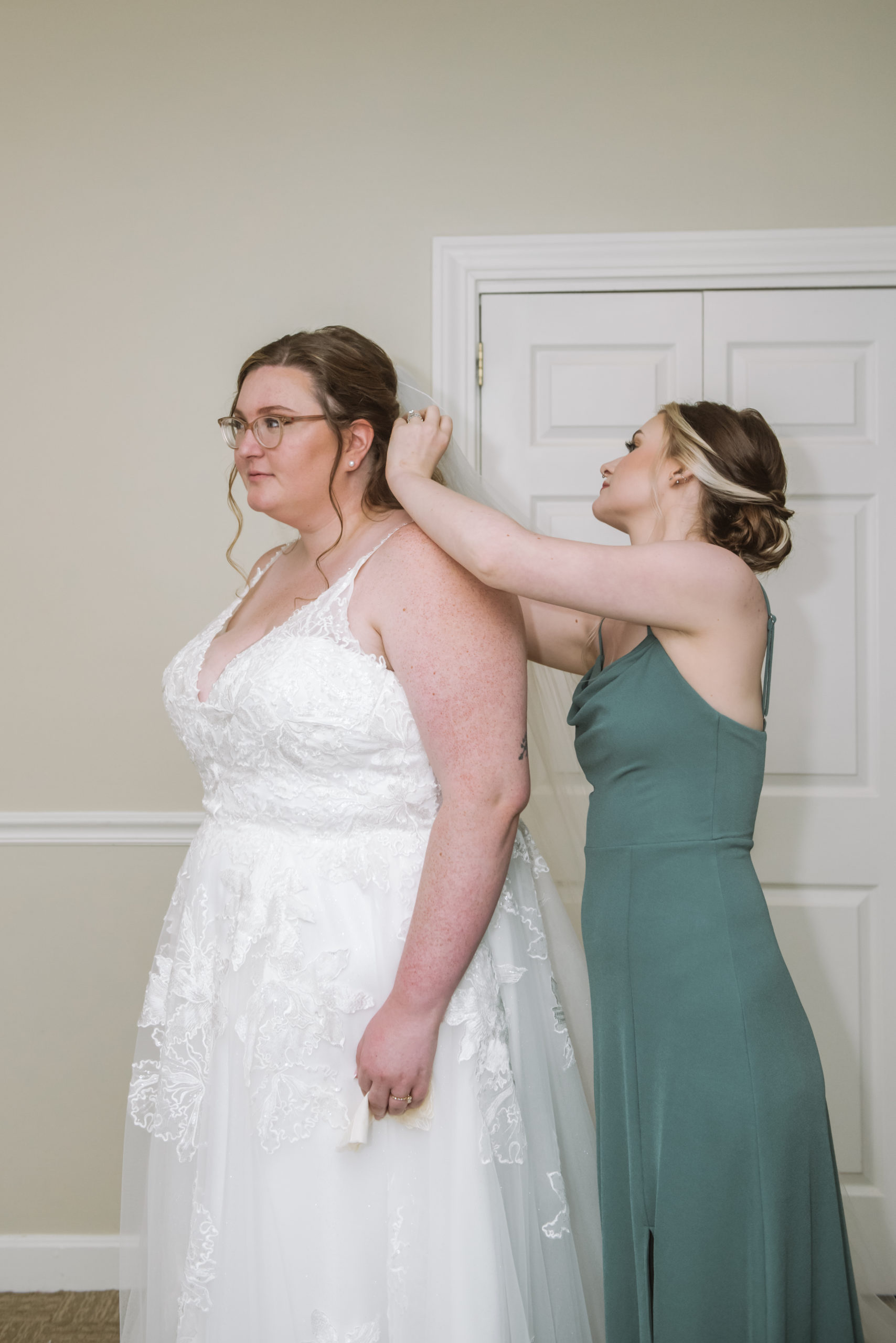 Maid of honor adjusting the bride's veil in a hotel room. The bride is dressed in her white lace long gown and the maid of honor is in her sage gown.