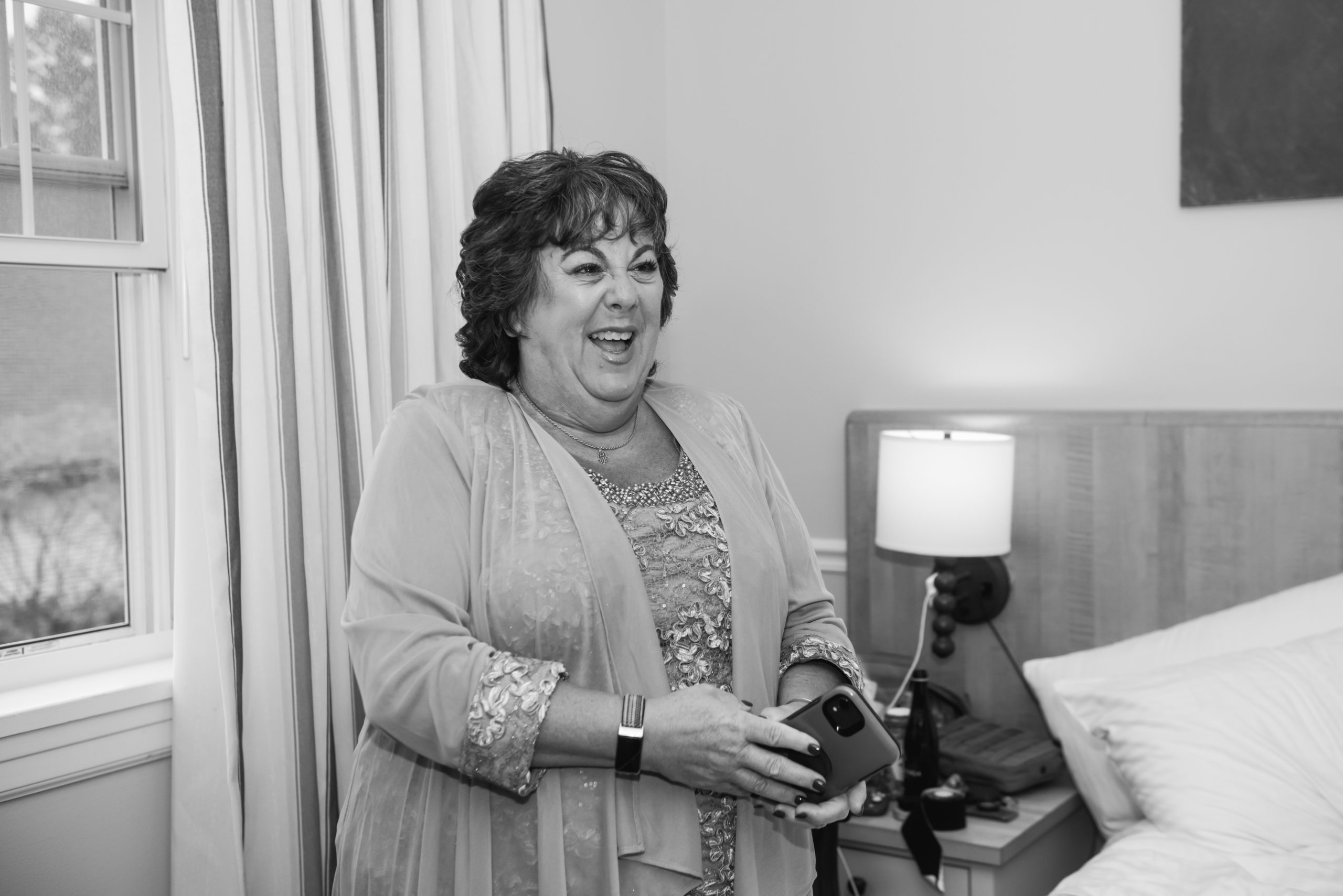 Black and white photograph of the mother of the bride smiling open-mouthed looking straight ahead, off camera. She is dressed for the wedding. She is standing in a hotel room.