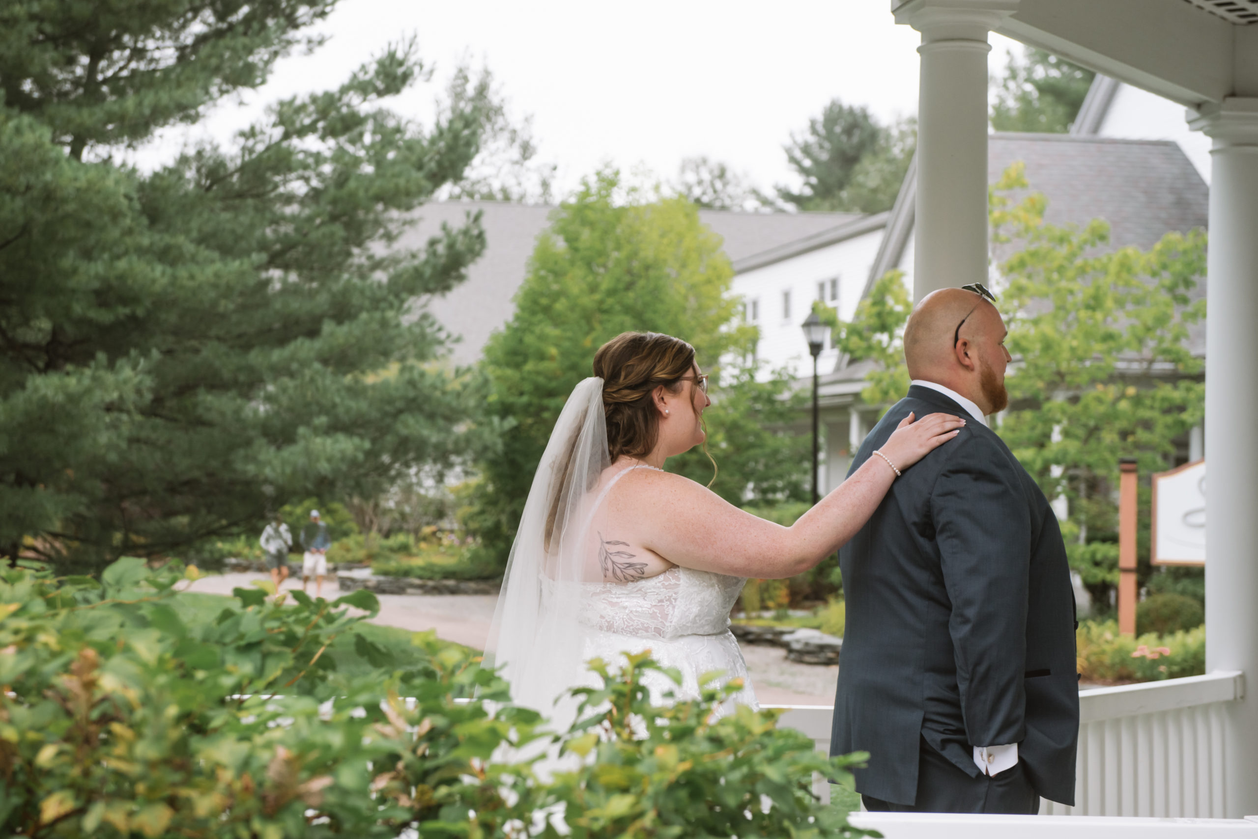 Bride standing behind her groom with her right hand on his right shoulder about to have the first look. His sunglasses are atop his head. They are standing in a covered walkway with bushes/greenery in the foreground and trees in the background. She is wearing a long white lace gown with a long veil and he is wearing a blue suit. 