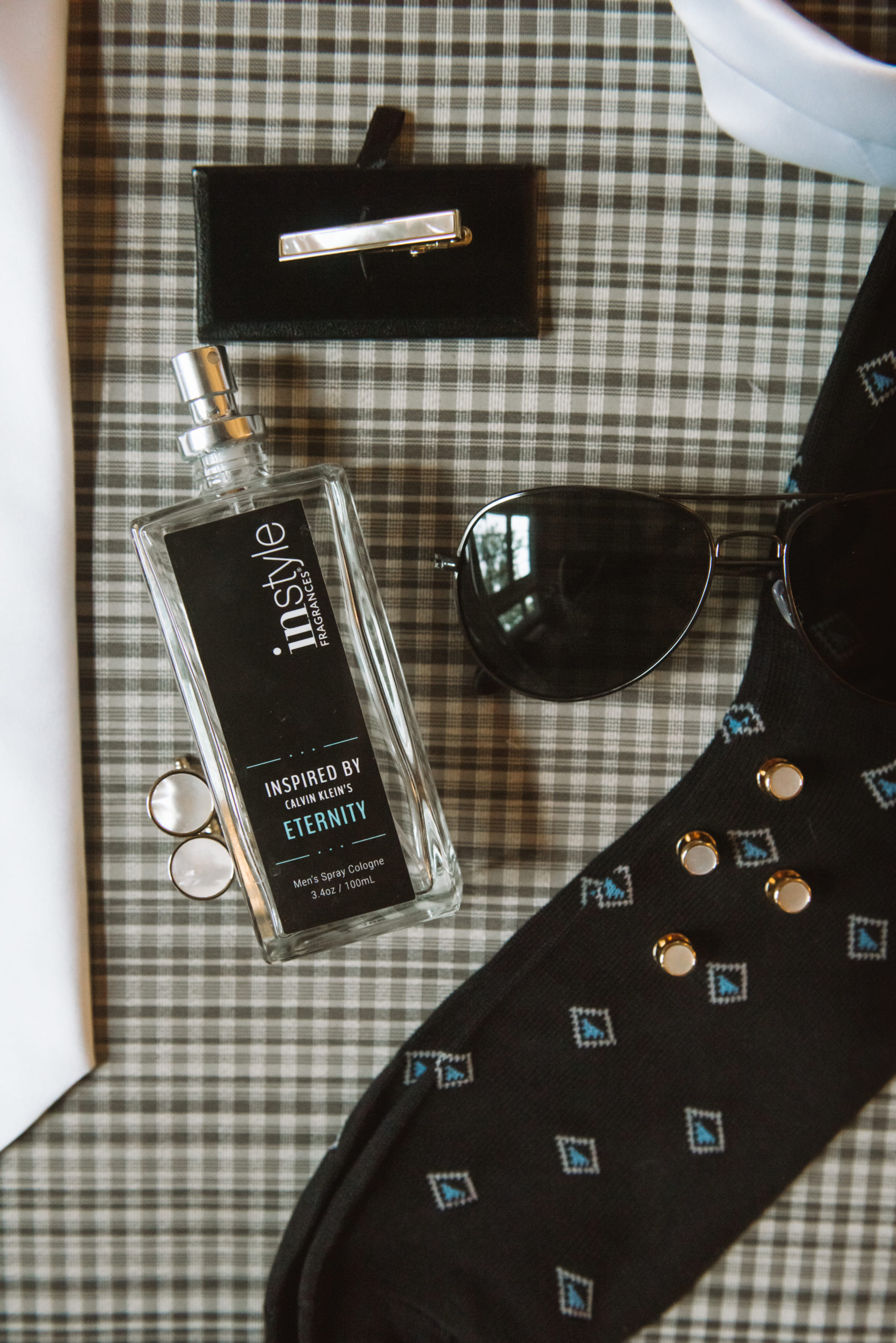 Close up detail of the groom's details including a bottle of cologne, printed socks, Ray Ban aviator sunglasses, white tie, and matching tie clip and cufflinks. Everything is set atop a green, black, and white gingham ottoman.
