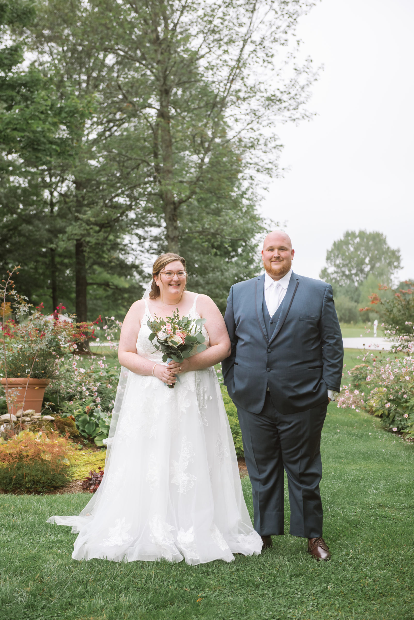 Bride and groom standing side by side in a garden. They are both standing facing the camera and have soft smiles. She is holding her bouquet in front of her. He has both hands in his pockets.  She is wearing a long white lace gown with a long veil and he is wearing a blue suit with a white tie. 