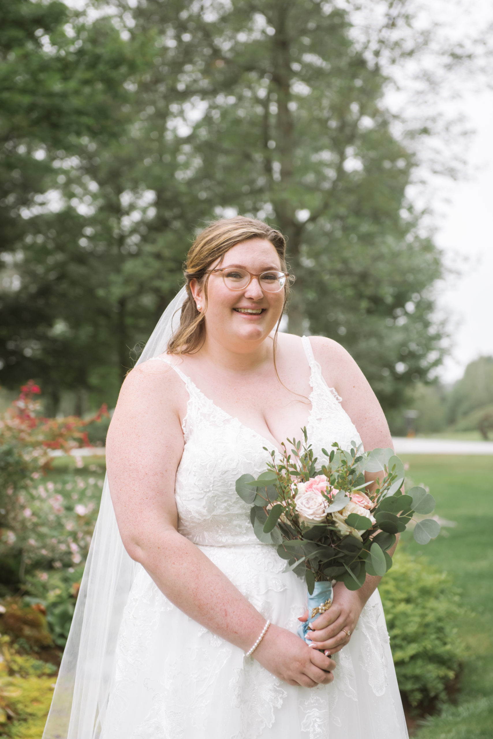 Torso portrait of the bride standing in a garden. She is holding her bouquet in front of her. She is wearing a long white lace gown with a long veil. She is smiling straight to camera and is wearing glasses.
