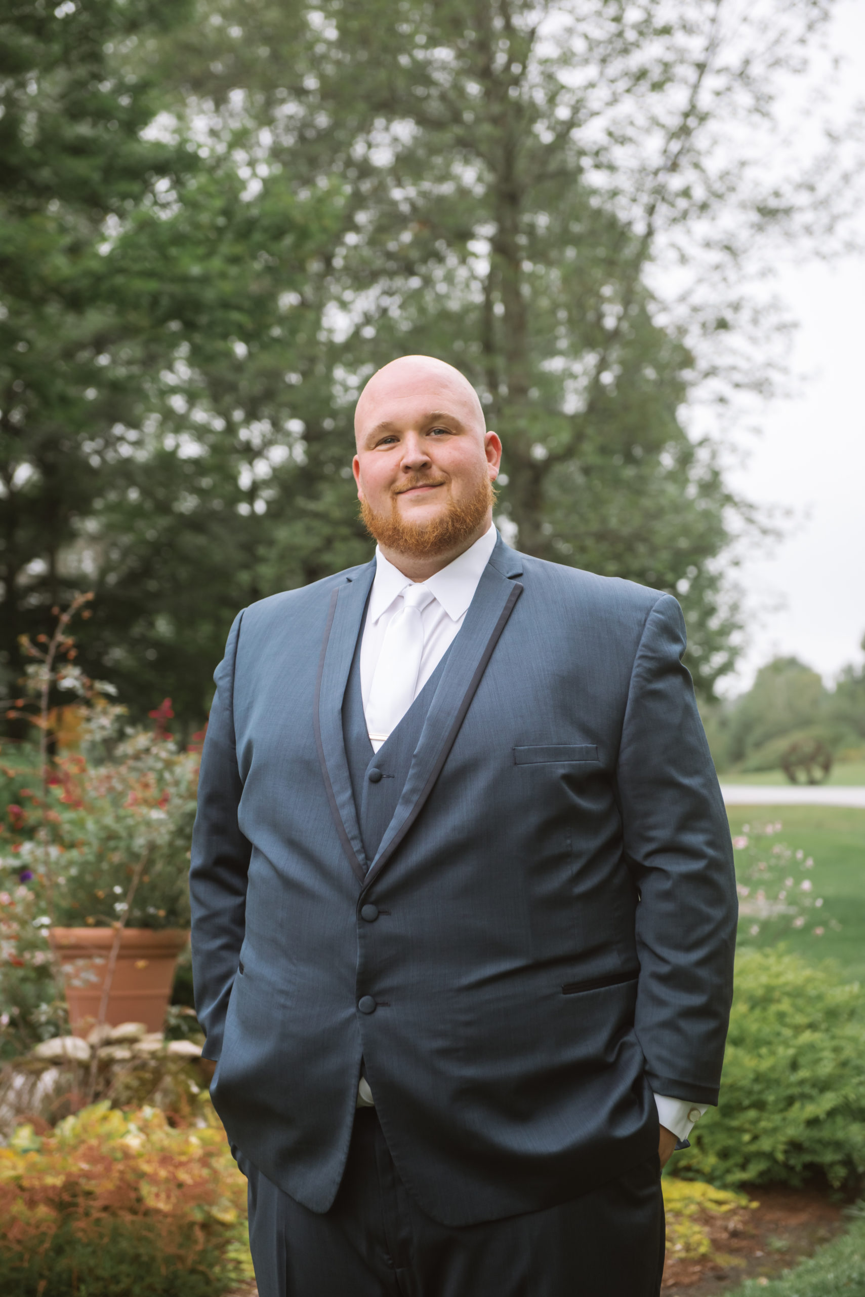 Torso portrait of the groom standing in a garden facing the camera and looking directly at the camera. He has a soft smile on his face. He is wearing a blue suit with a white tie. Both of his hands are in his pockets.